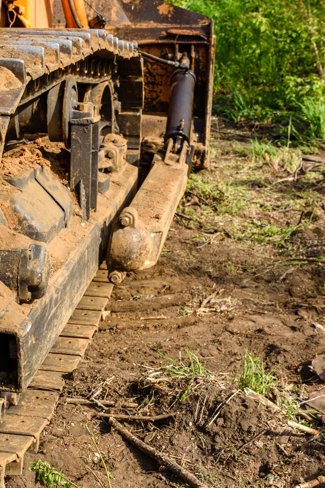 Close-up of a bulldozer's metal tracks and hydraulic system on a dirt field with green foliage in the background.