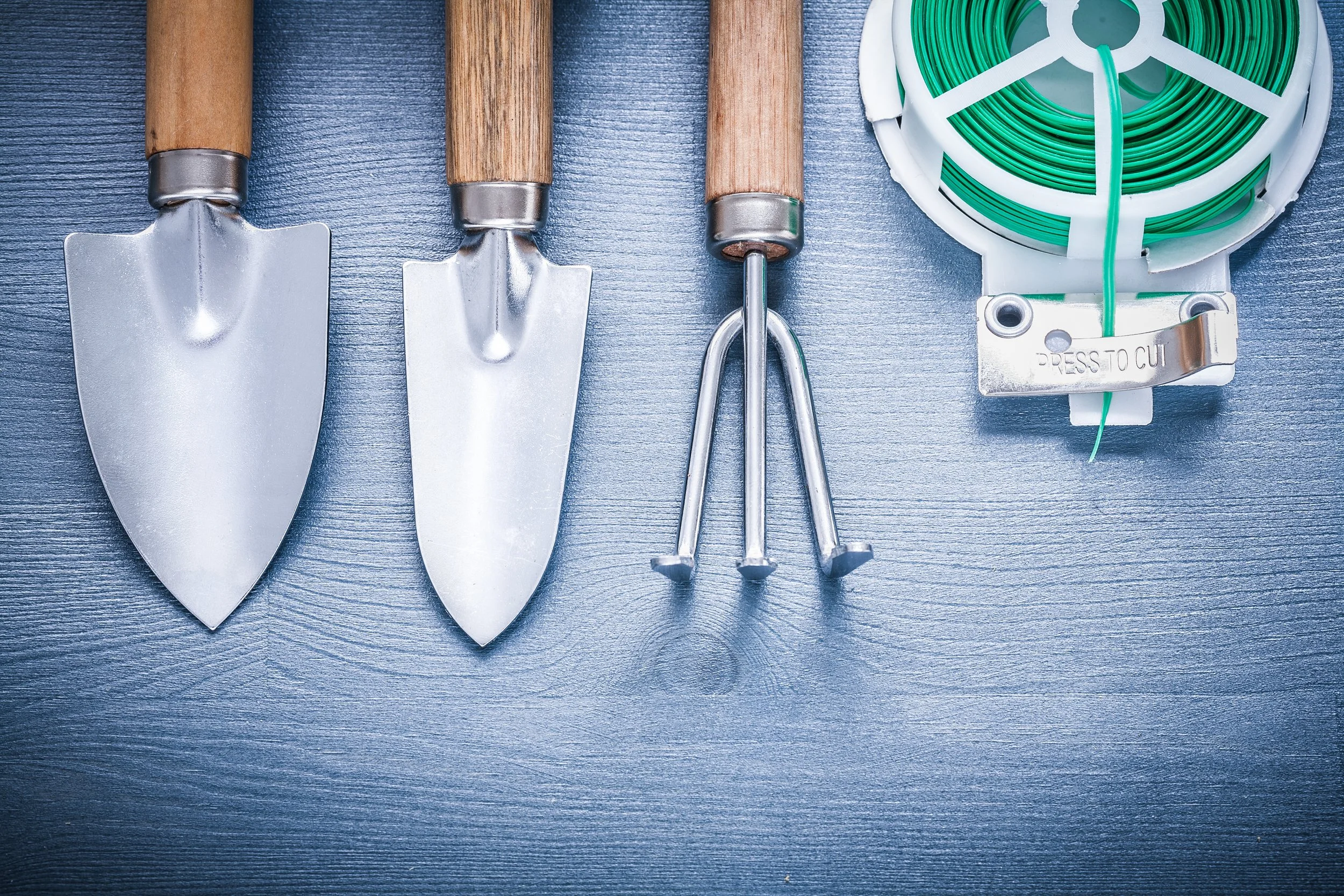 Four gardening tools with wooden handles and metal heads, and a plastic reel of green plant tie wrap on a dark gray textured surface.
