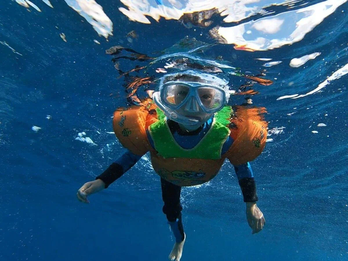 Child snorkeling underwater wearing a mask, snorkel, and orange life vest, swimming in blue water.