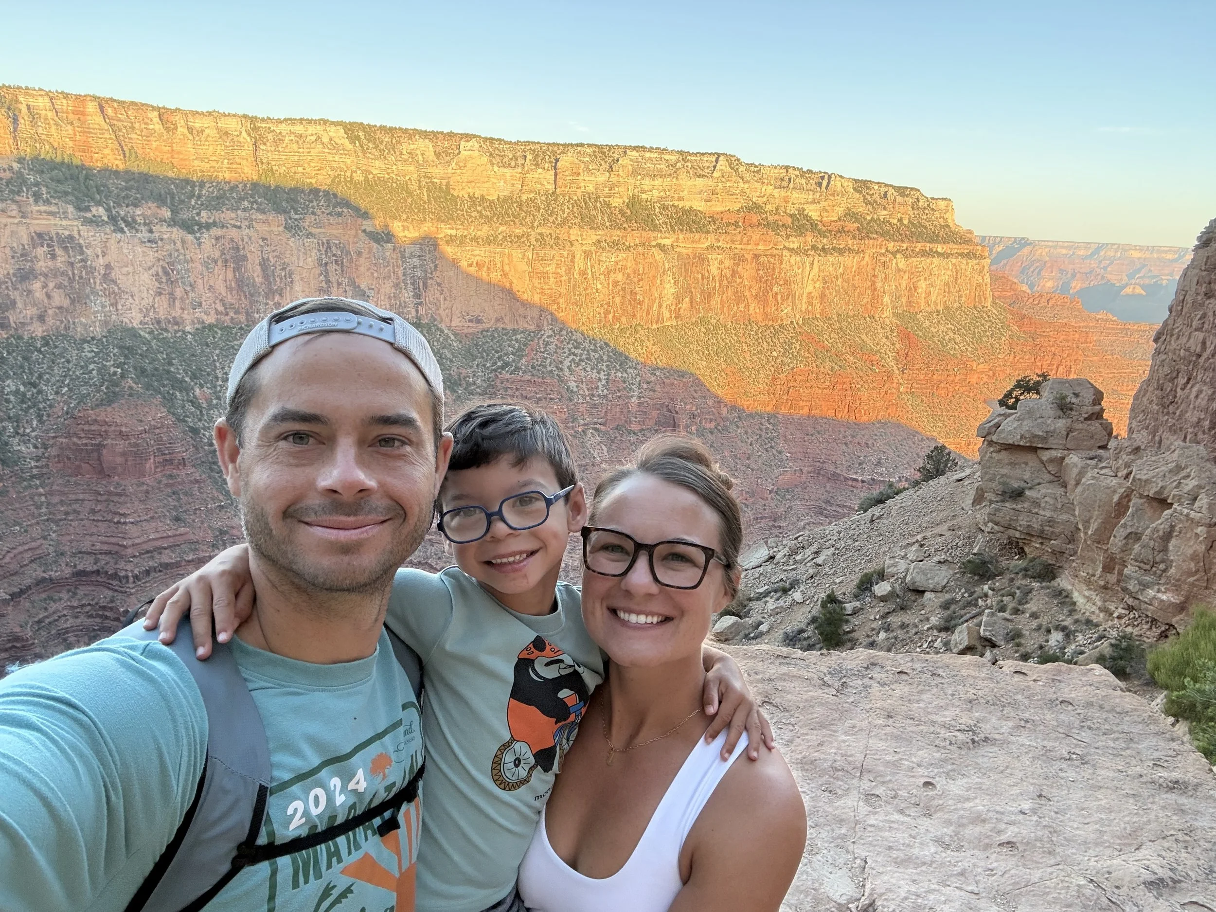 A group of four people smiling at the camera during a visit to the Grand Canyon, with the canyon's layered red rock formations and cliffs visible in the background at sunset.