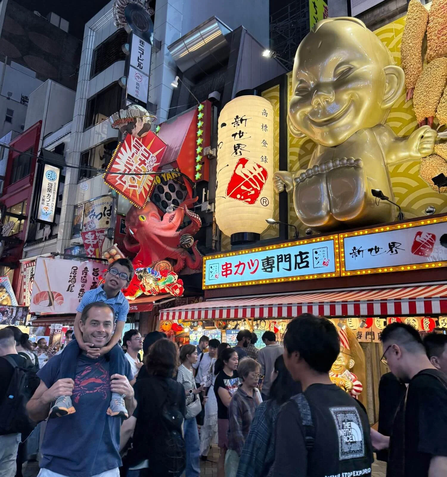 Son on dad's shoulders on Dotonbori street in Osaka Japan with baby buddha in the background lit up at night.