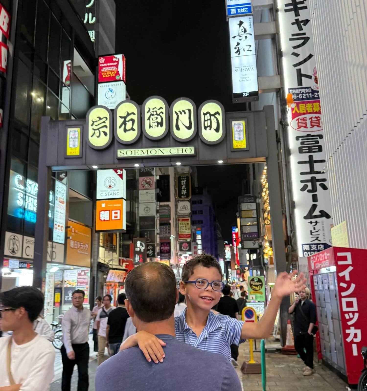 A father carrying his smiling, waving son on Dotonbori Street in Osaka, Japan, with neon signs lit up behind them.
