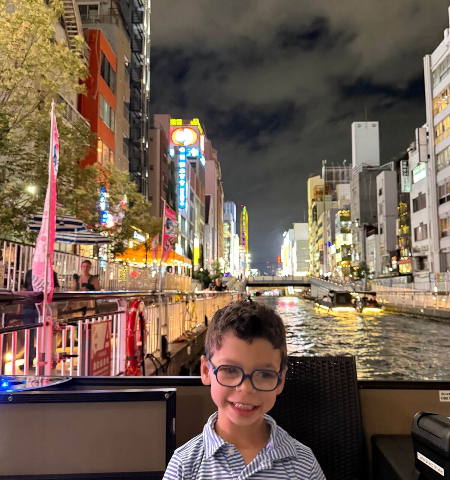 A young boy on a canal boat ride in Dotonbori, Osaka, Japan, with neon lights reflecting on the water behind him.