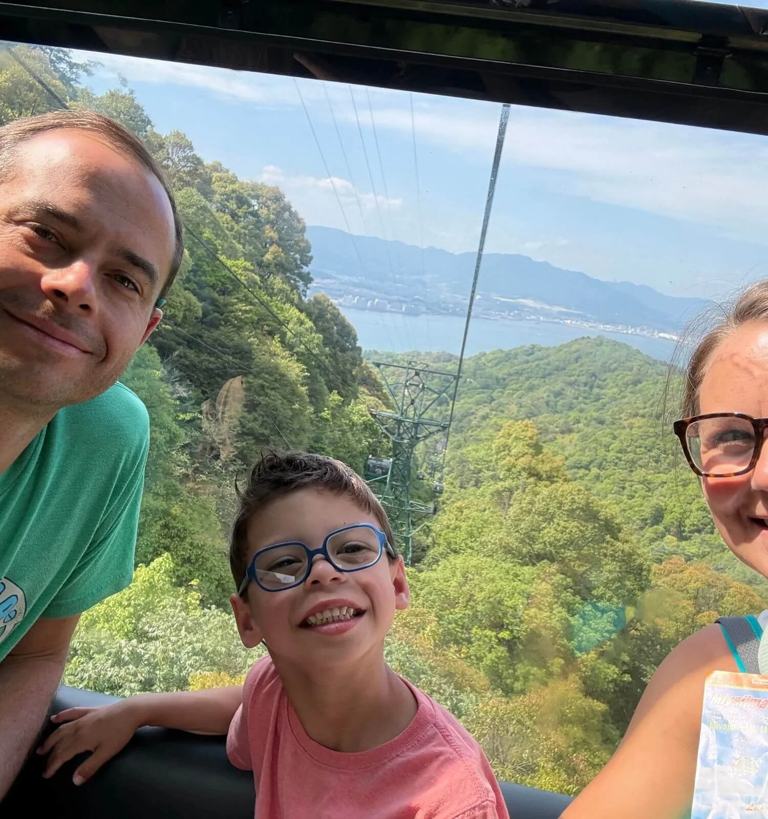 Family selfie inside the Miyajima Ropeway with views of the water and Hiroshima in the background.