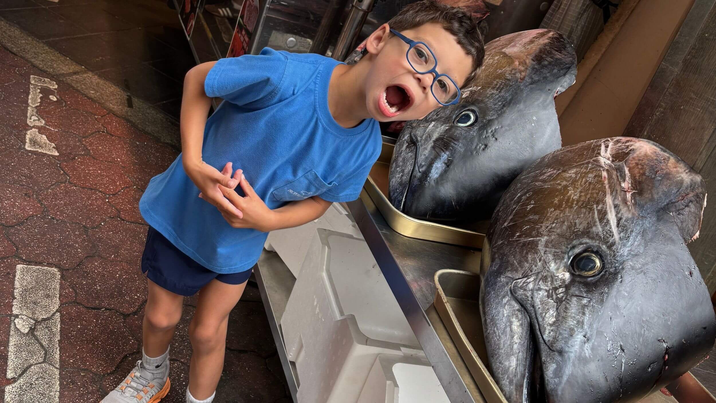 Young boy making a shocked expression next to large tuna heads at Tsukiji Market in Tokyo, Japan