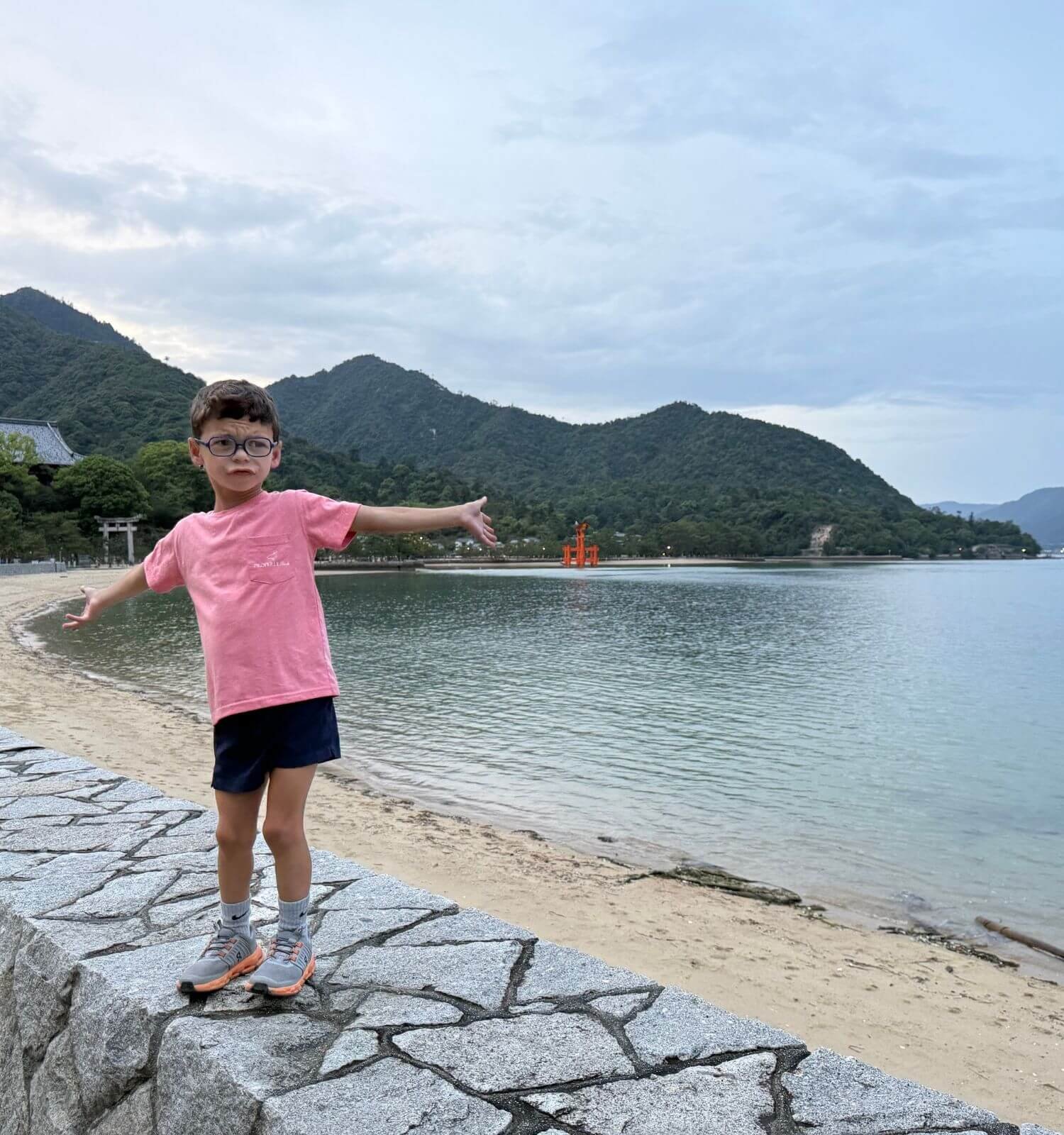 A young boy making a funny face with arms spread wide while standing on a rock wall in Miyajima, Japan, with Itsukushima Shrine visible behind him.