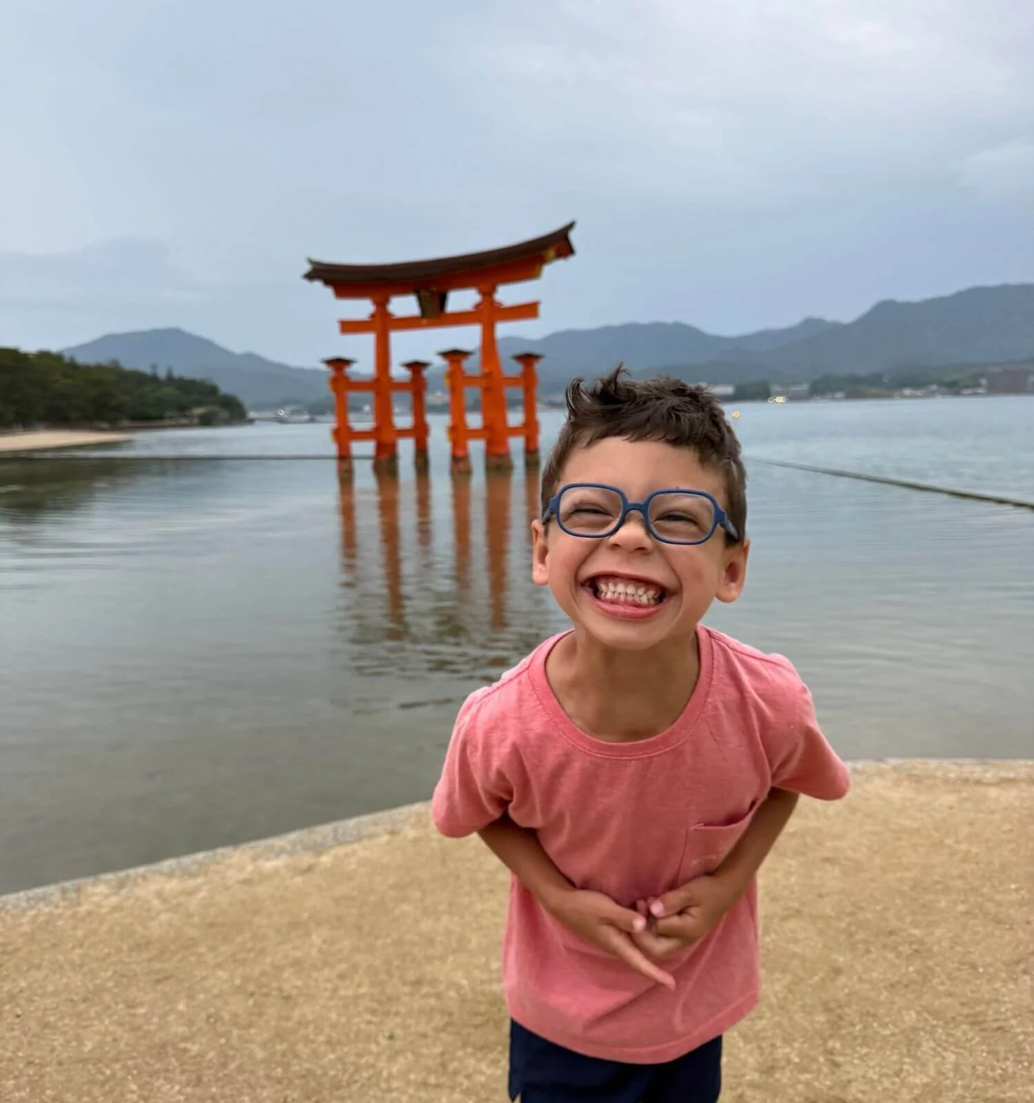 A young boy smiling big with the floating torii gate visible behind him in Miyajima, Japan.