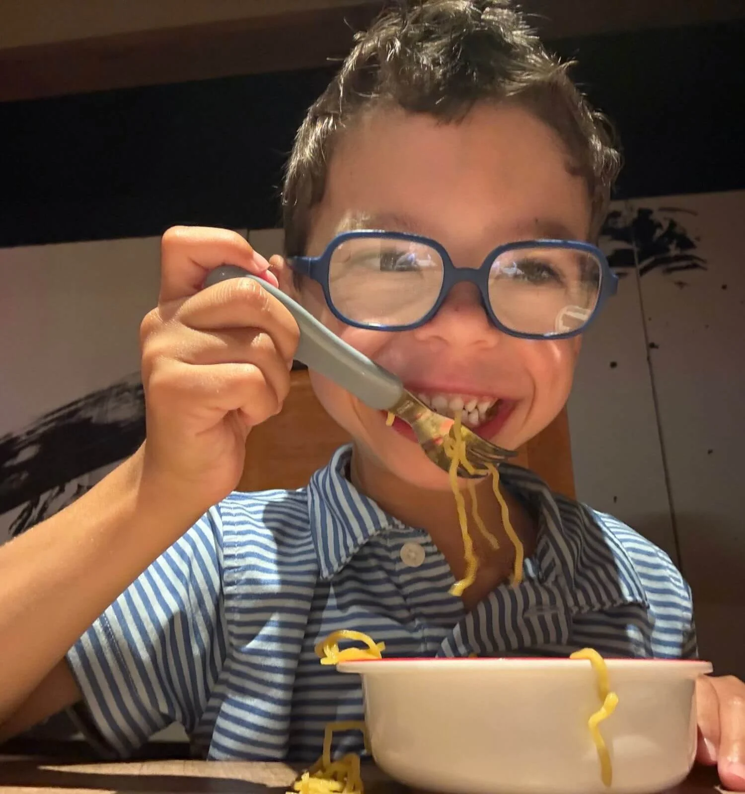A young boy smiling and eating ramen in Osaka, Japan, his glasses fogged up from the steam of the noodles.