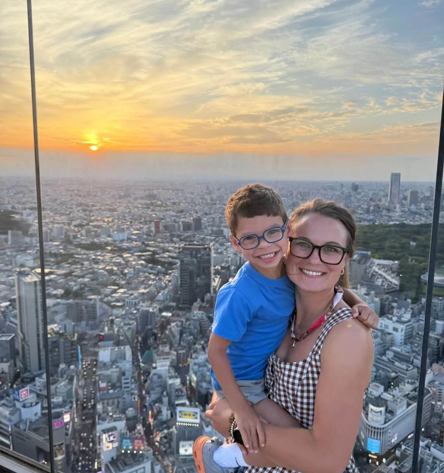 A mother and son smiling at Shibuya Sky observation deck in Tokyo, Japan, with a colorful sunset over the city skyline behind them.
