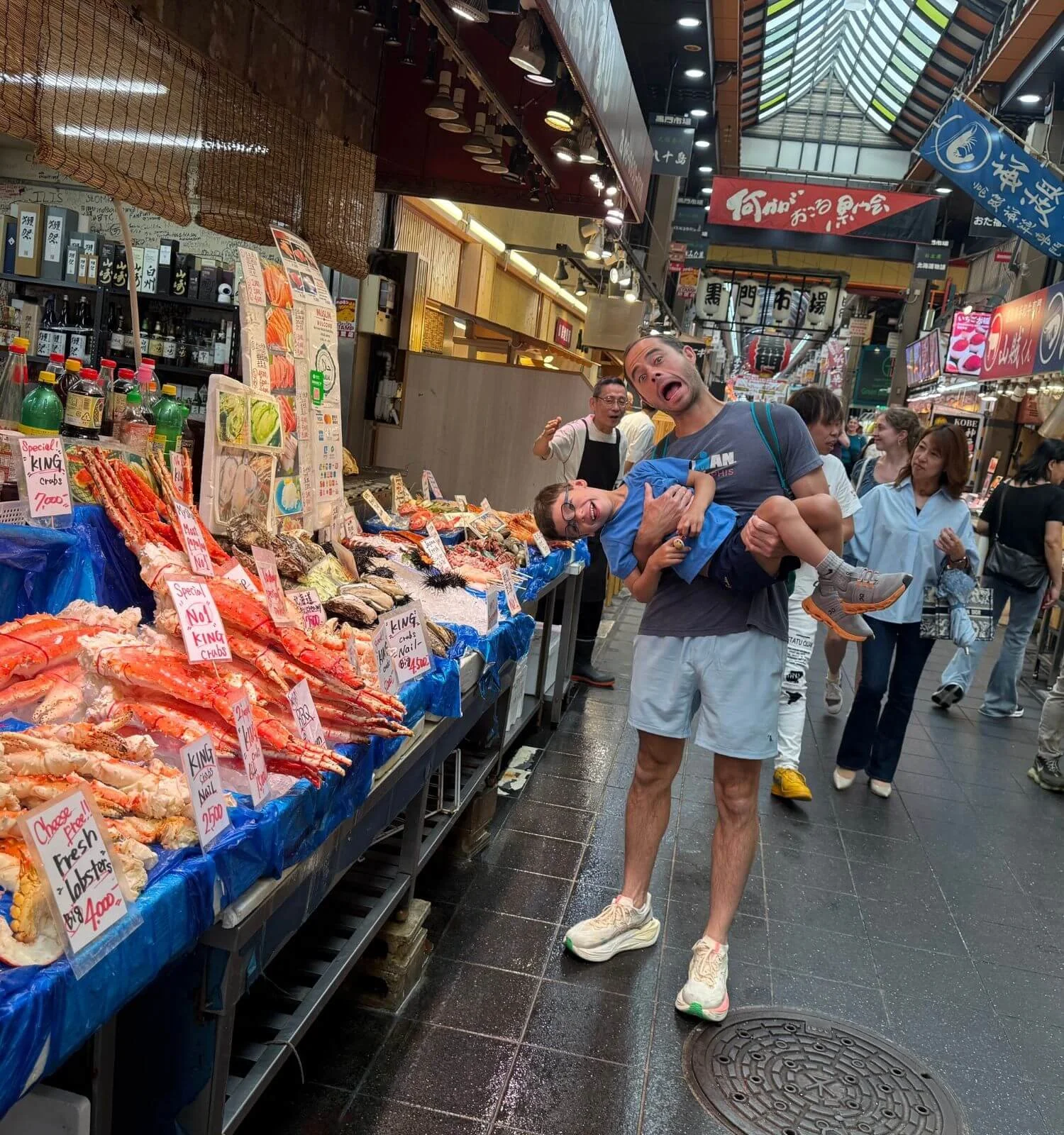 A father pretending to put his son's head into a display of seafood at Kuromon Market in Osaka, Japan, both making silly faces.