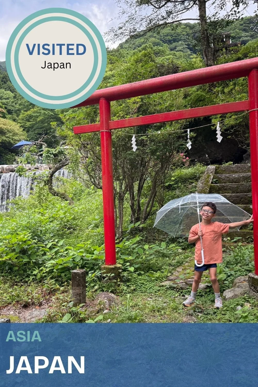 A young boy standing near a red torii gate with a clear umbrella, smiling in a lush green forest with a waterfall in the background in Japan.
