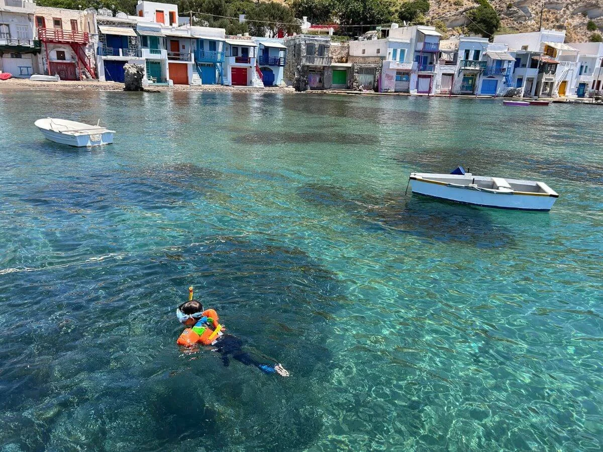A person snorkeling in clear blue water near colorful buildings by the shoreline, with small boats floating nearby located in Milos, Greece.