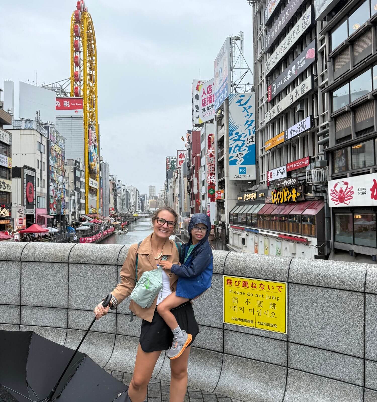 Child and parent standing on a bridge overlooking the Dotonbori Canal in Osaka, Japan, with the Ferris wheel and city skyline in the background.