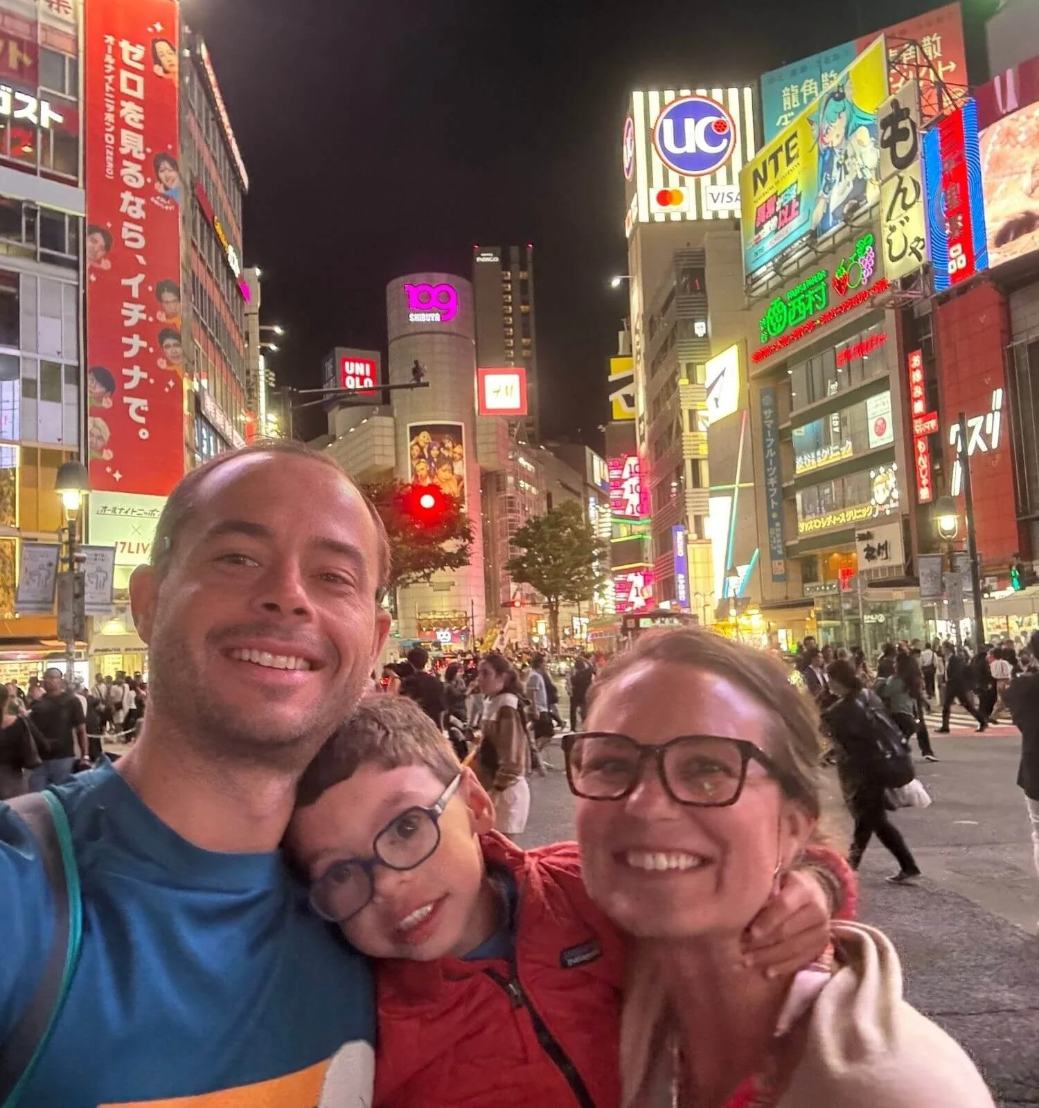 A family of three taking a selfie at Shibuya Crossing in Tokyo, Japan, one of the world's busiest pedestrian intersections.