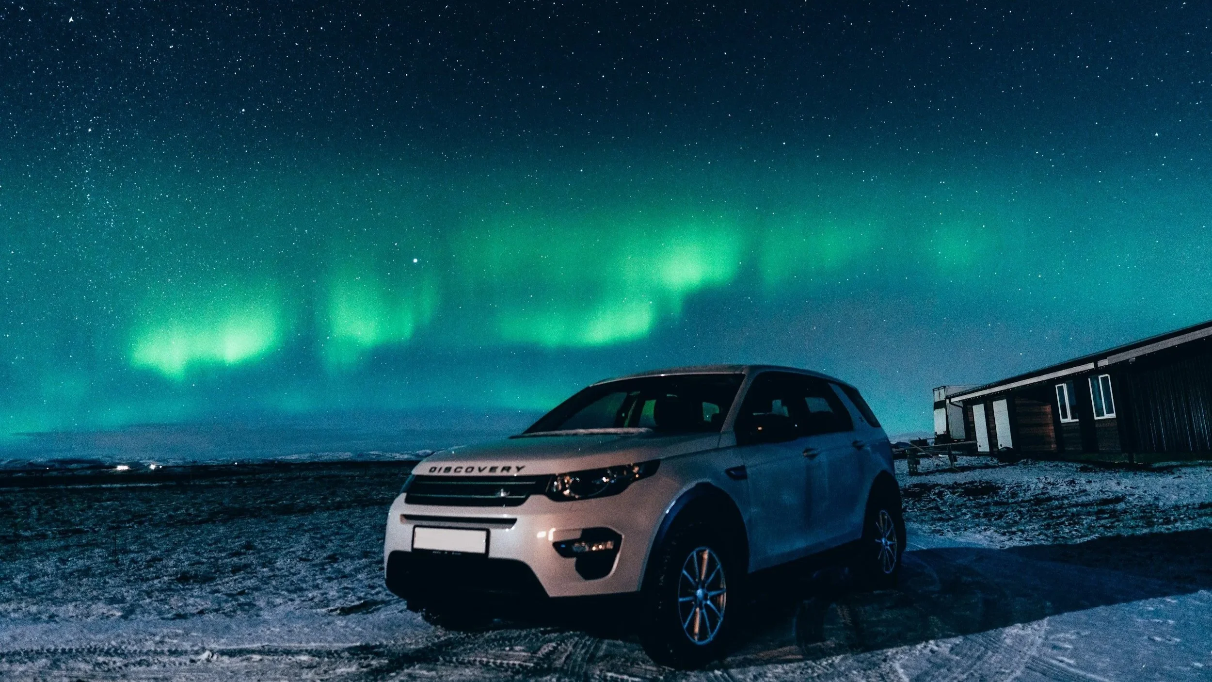 Bildet viser en hvit bil parkert på en snødekt overflate under en klar natthimmel med nordlys. Nordlyset er grønt og danser over stjerneklar himmel.