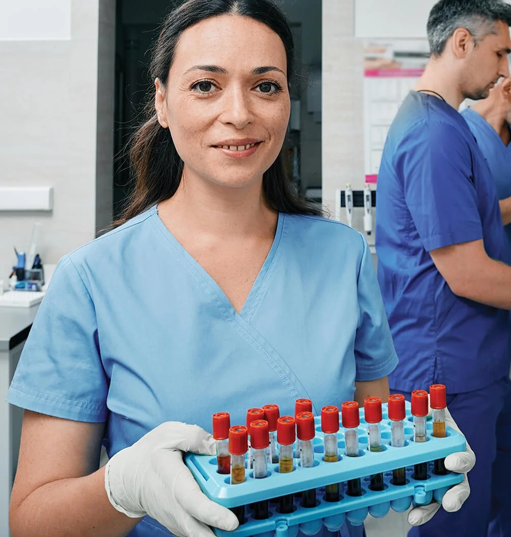 Healthcare laboratory technician holding a tray of blood sample tubes while working in a medical lab.