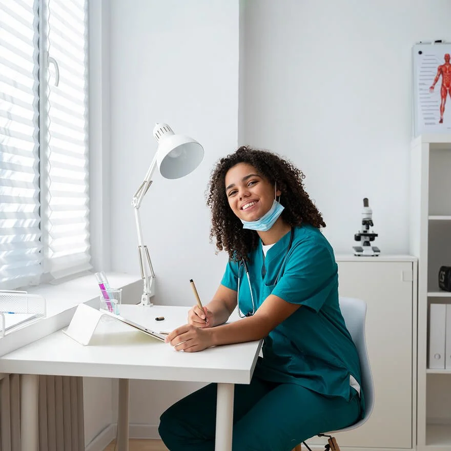 Healthcare professional sitting at a desk with a laptop and medical mask, smiling in a clinical office.