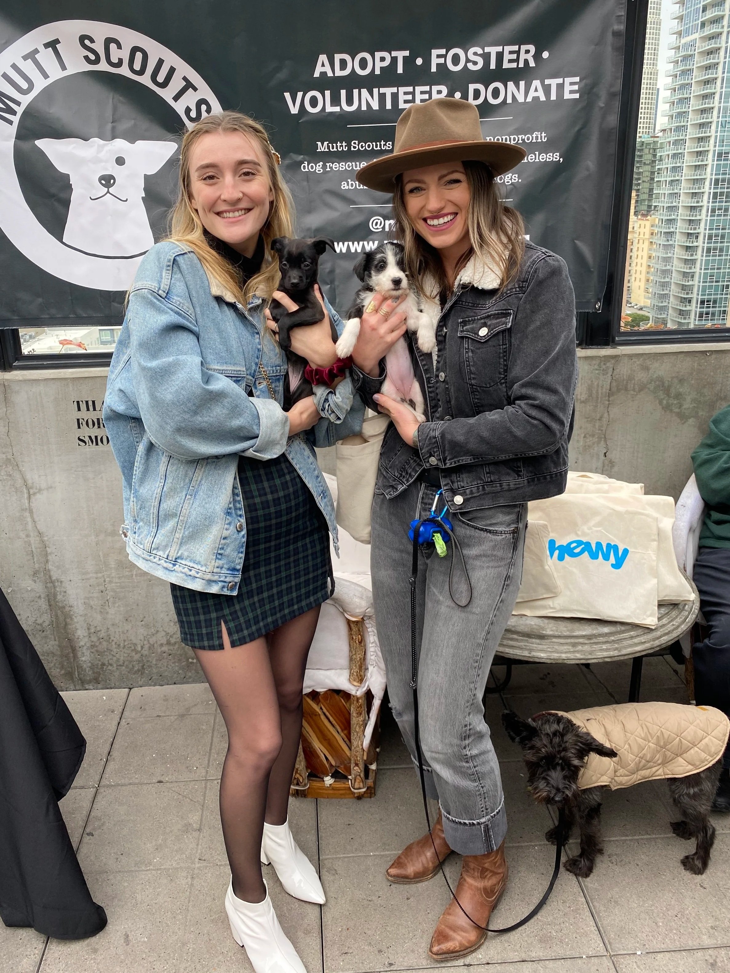 Two women holding puppies at an outdoor event with a banner in the background that reads 'Adopt Foster Volunteer Donate' for a dog rescue organization. The woman on the left is wearing a denim jacket and a plaid skirt, while the woman on the right is wearing a black jacket, jeans, and a wide-brimmed hat. There is a black dog with a beige coat on the ground near the woman on the right.