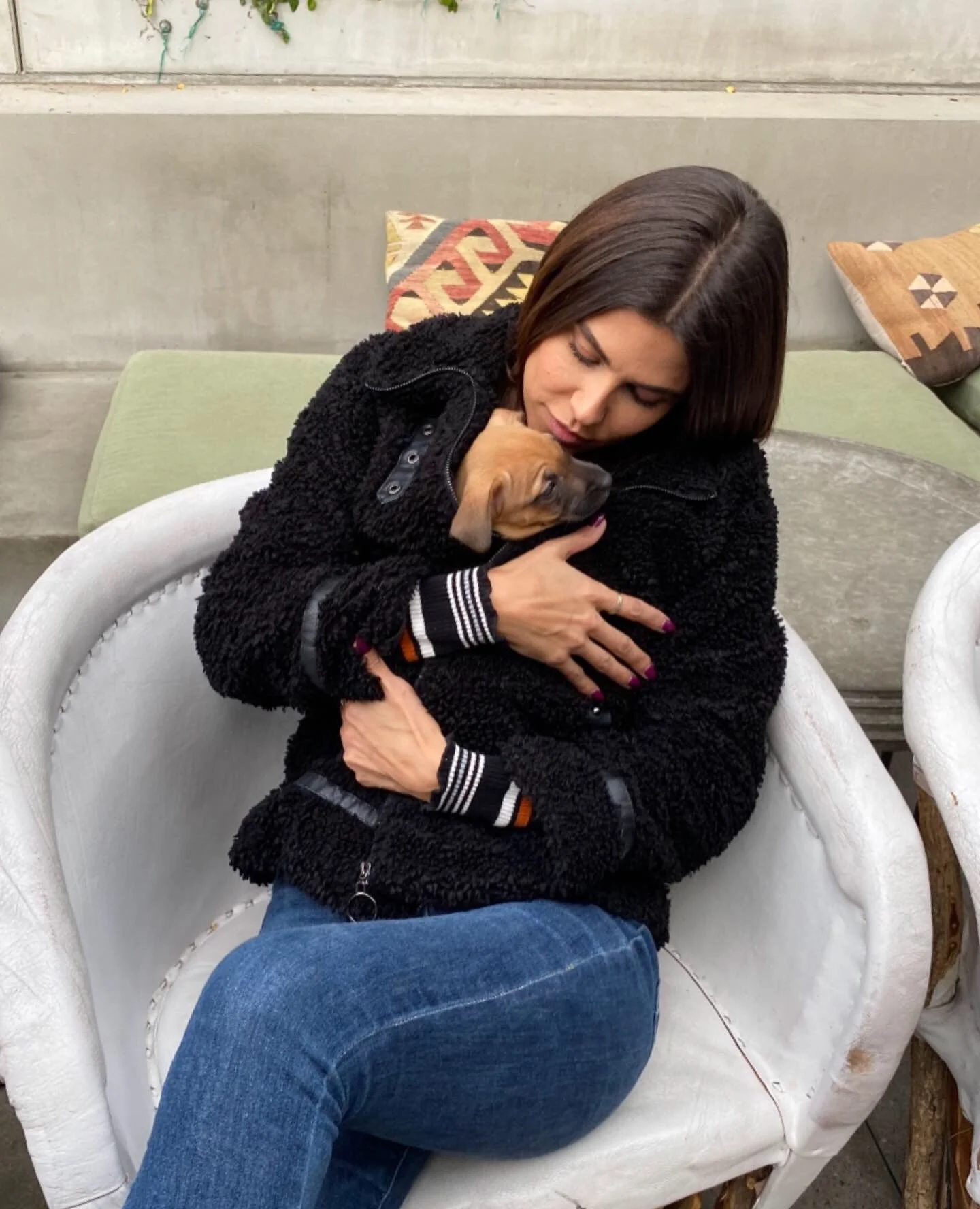 A woman with brown hair wearing a black jacket and blue jeans sitting on a white chair, hugging a small brown puppy, with colorful pillows behind her.