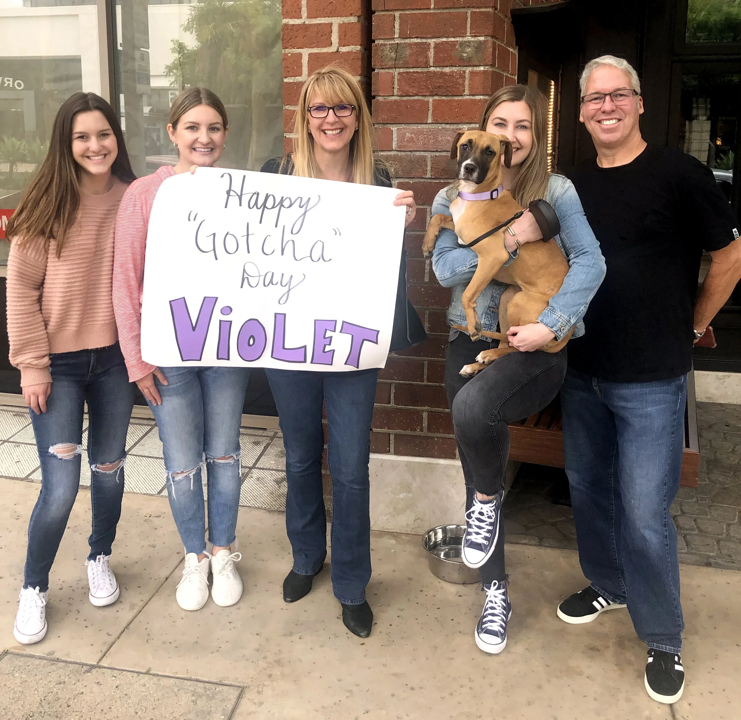 Group of five people, four women and one man, standing outside near a brick wall, holding a sign that reads Happy Gotcha Day Violet. One woman is holding a brown dog with a purple collar. All are smiling and dressed casually.