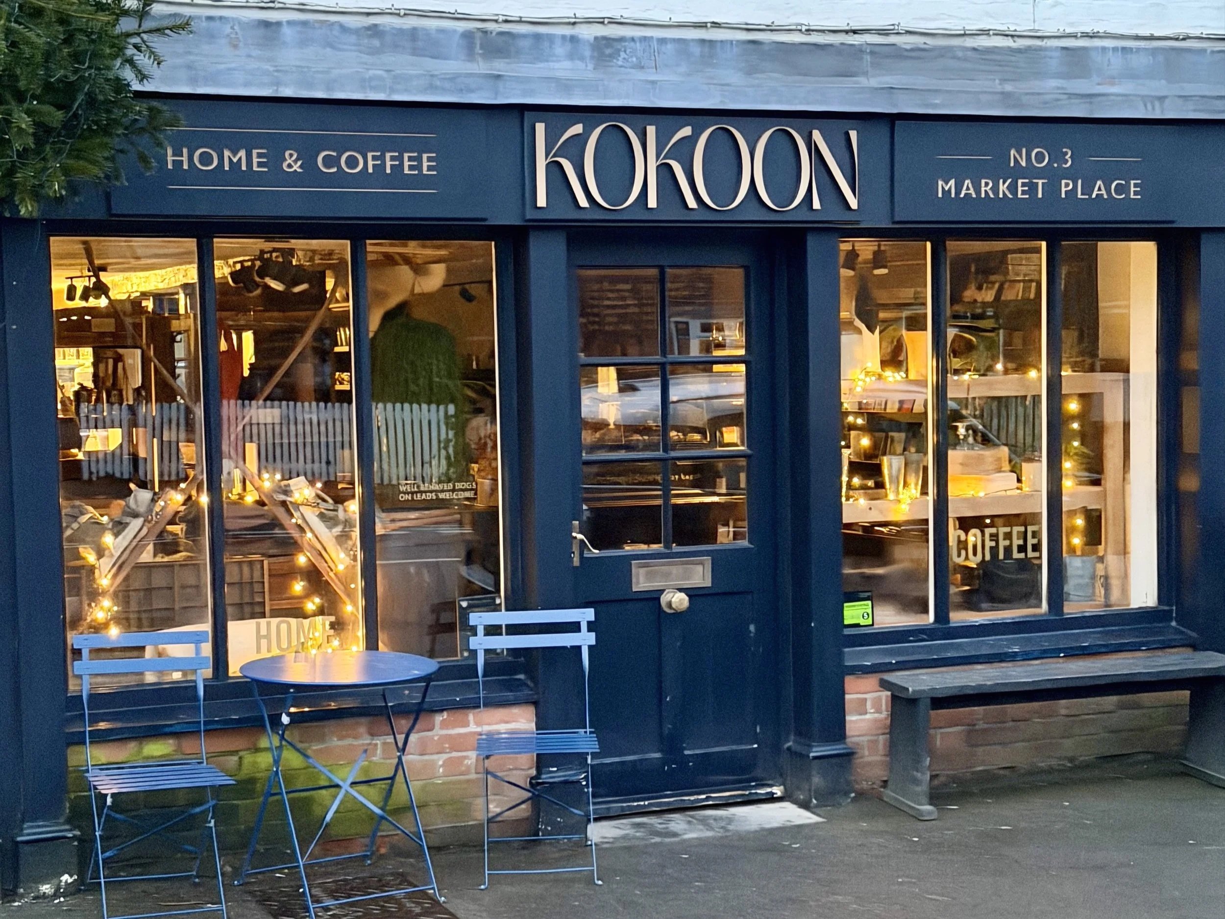 Front view of a coffee shop with blue exterior, large front windows, and outdoor seating including a small table and two chairs, decorated with string lights inside.