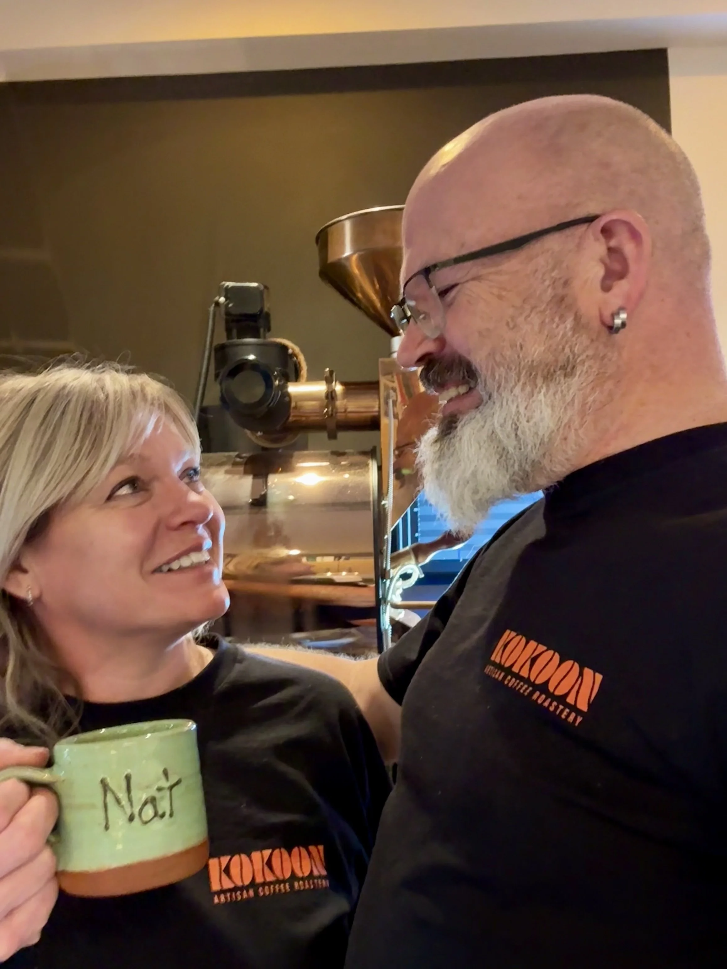A woman holding a green coffee mug labeled 'Nat', looking up at a man with glasses, a beard, and earrings, smiling at each other inside a coffee shop.