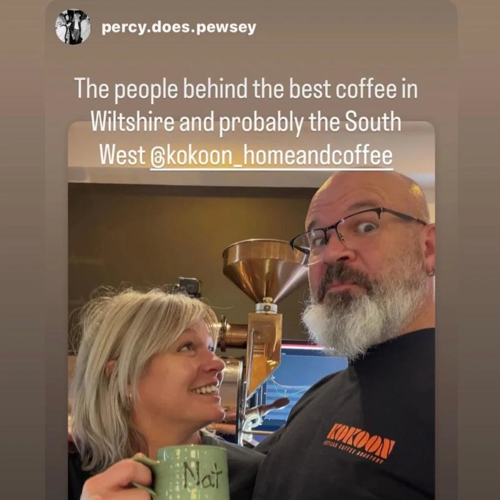A woman and a man are posing for a selfie indoors in front of a coffee roasting machine. The woman is smiling and holding a green mug with the word 'Nat' written on it. The man has a surprised expression, with glasses and a beard, wearing a black shirt with an orange logo that reads 'Kokoon.' The wall and ceiling are beige, and the coffee roasting equipment is visible in the background.