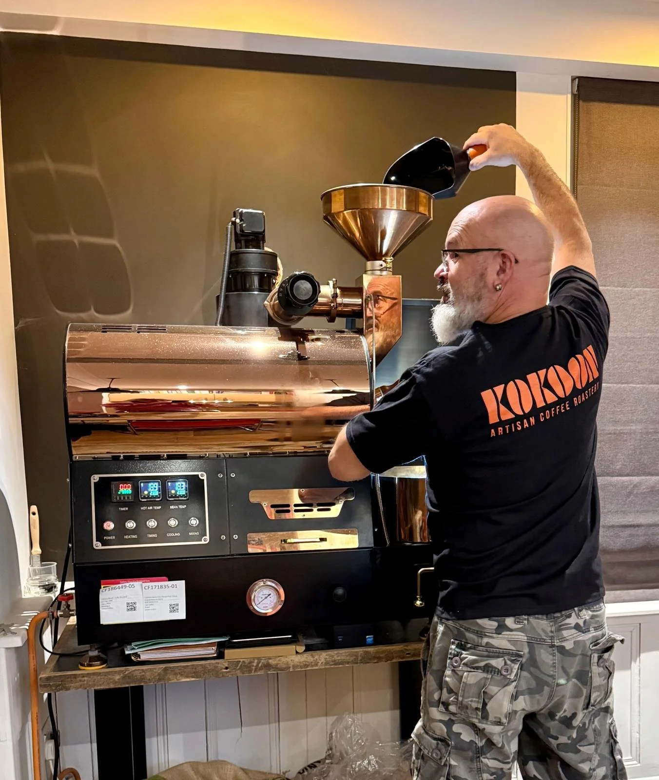 A man with a bald head and gray beard wearing glasses and a black T-shirt with orange text 'KOKOOM ARTISAN COFFEE ROASTER' is operating a large, shiny, copper-colored coffee roasting machine.