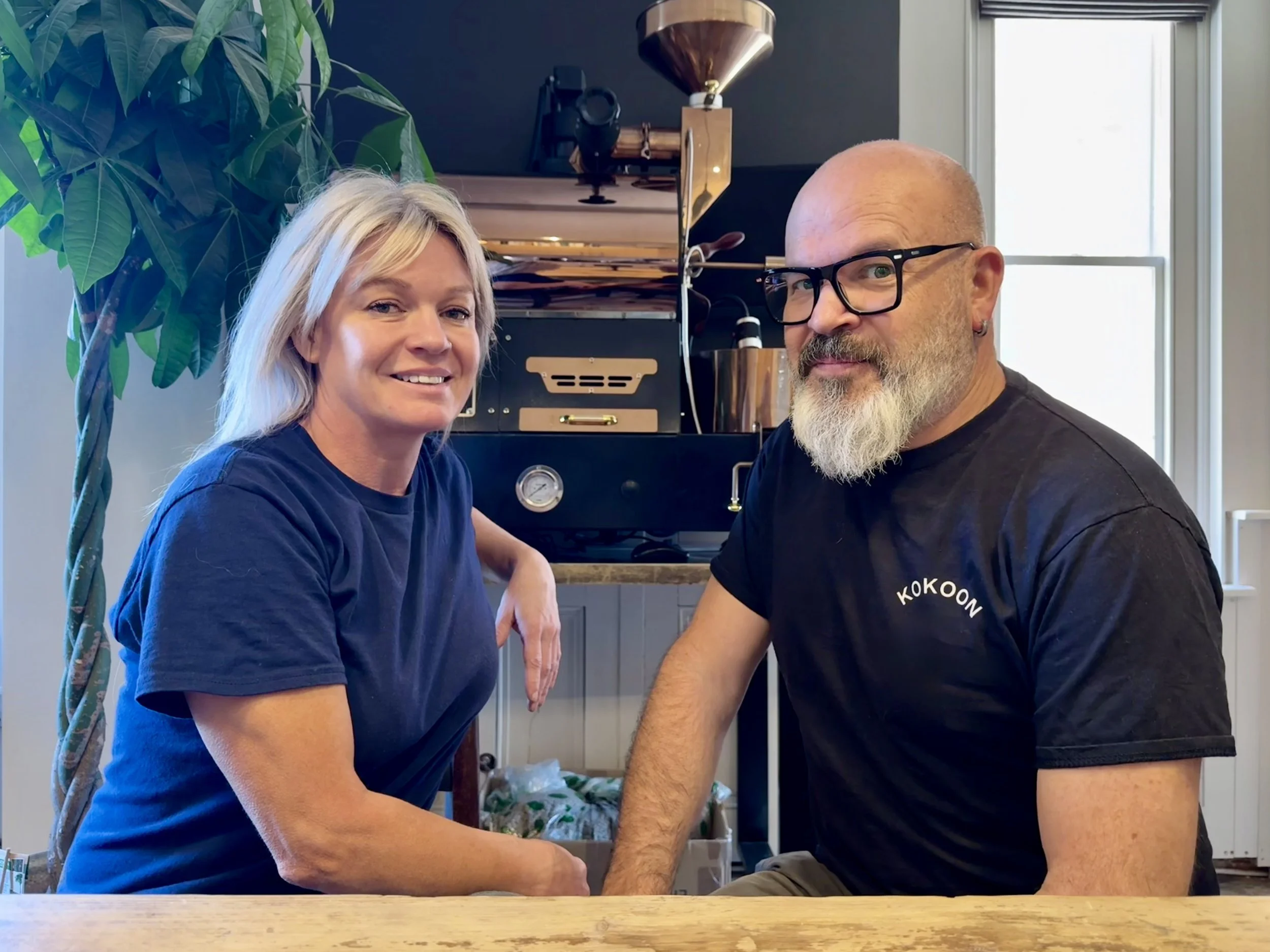 A woman and a man sit close together at a wooden table in a cozy indoor setting, smiling at the camera, with a large plant, some coffee roasting equipment, and a window in the background.