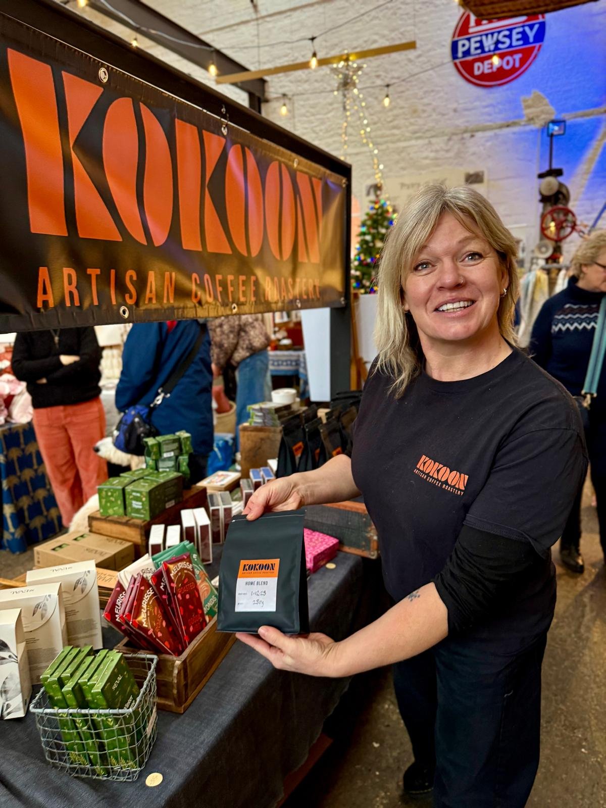 A woman with blonde hair smiling and holding a bag of coffee at a market stall named KOKOON. The stall offers artisan coffee and has various coffee products displayed on the table. There is a Christmas tree and holiday decorations in the background.