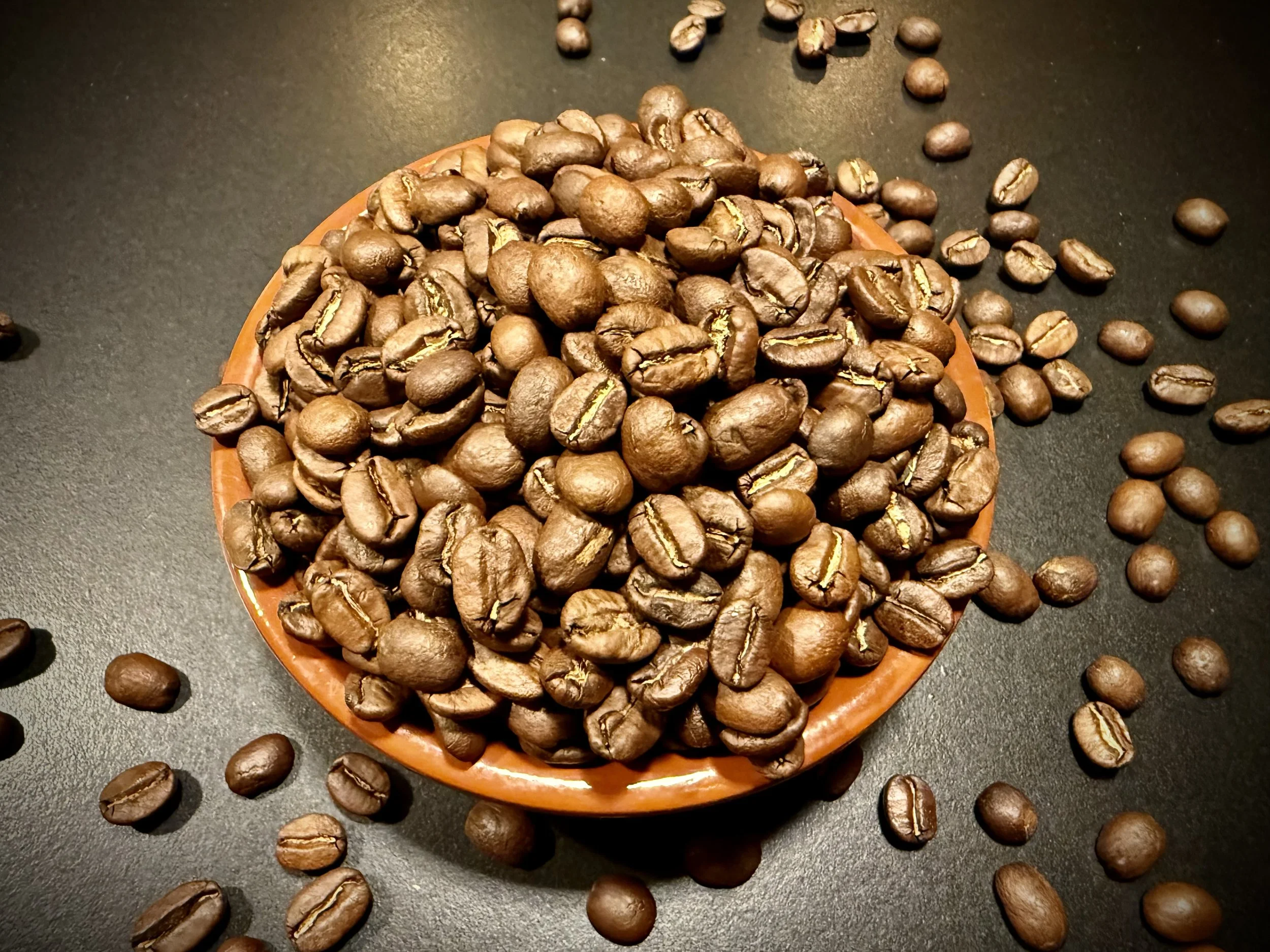 A bowl of roasted coffee beans on a dark surface with some beans spilled around.