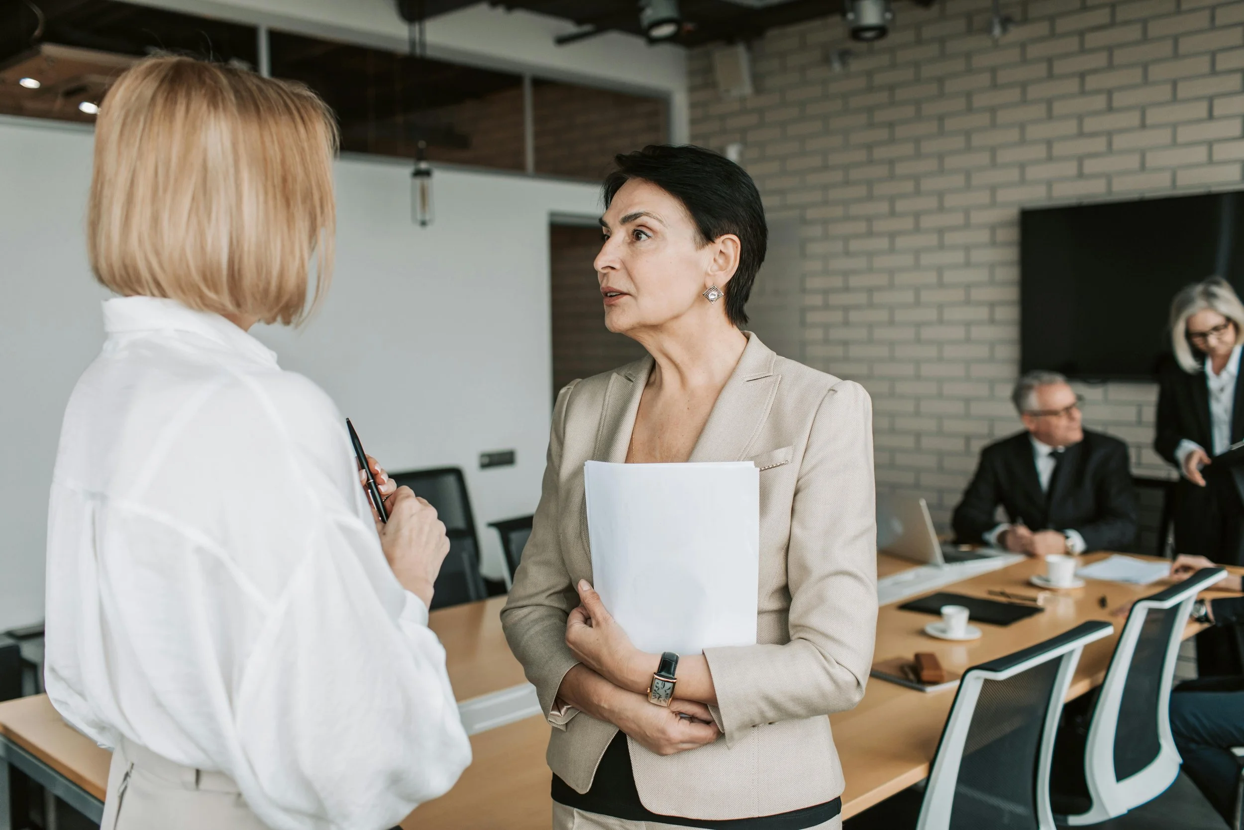 Two women engaged in a conversation in a modern office conference room. The woman on the right is dressed in a beige blazer, holding a white folder, and the woman on the left has blonde hair in a white blouse. In the background, three people are seated at a table with coffee cups and laptops, and a woman stands talking to another seated woman.
