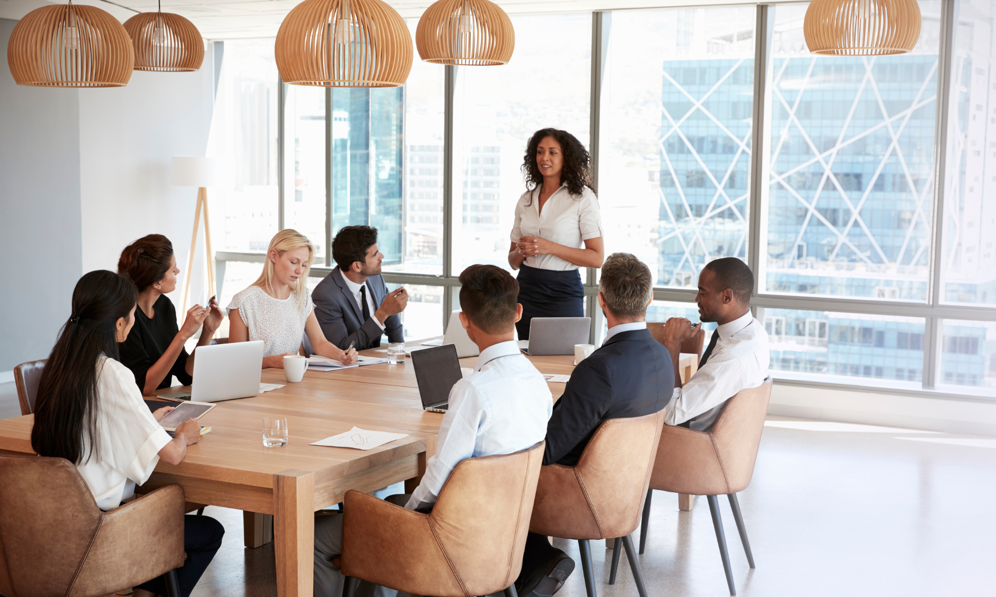 A woman standing and speaking to a group of seven people seated around a conference table in a modern office with large windows and a cityscape view.