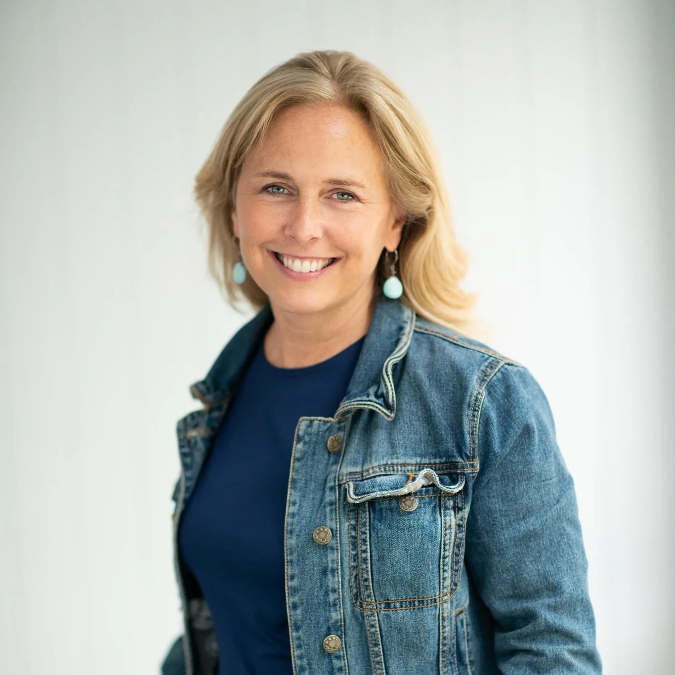 A woman with blonde hair smiling, wearing a navy blue shirt, denim jacket, and turquoise earrings, standing against a plain light background.