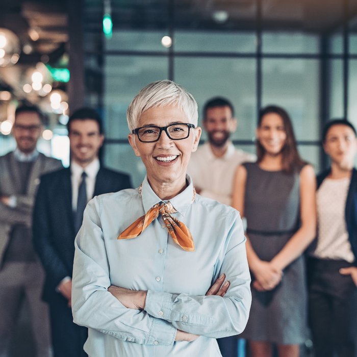 An older woman with short white hair, glasses, and a light blue shirt with an orange scarf stands smiling with arms crossed in front of a group of diverse young professionals in an office setting.