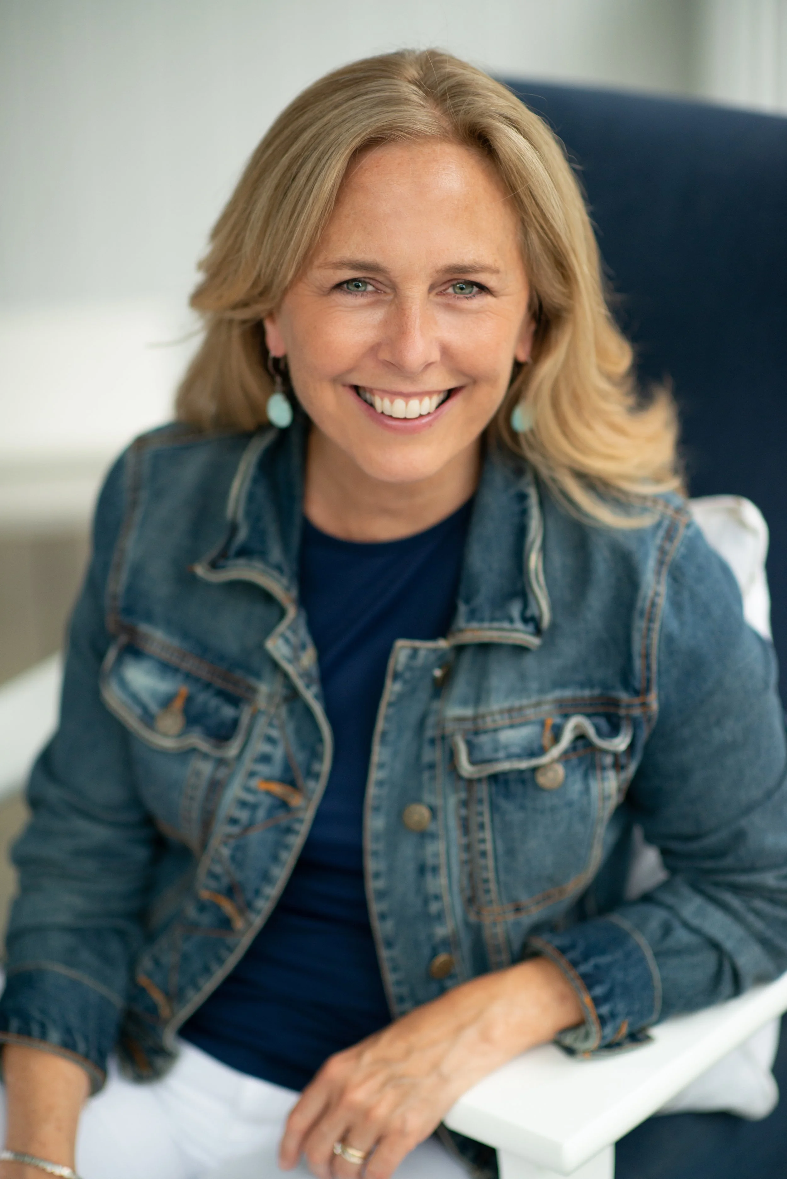 A woman with blonde hair wearing a denim jacket and turquoise earrings, smiling while sitting on a chair.