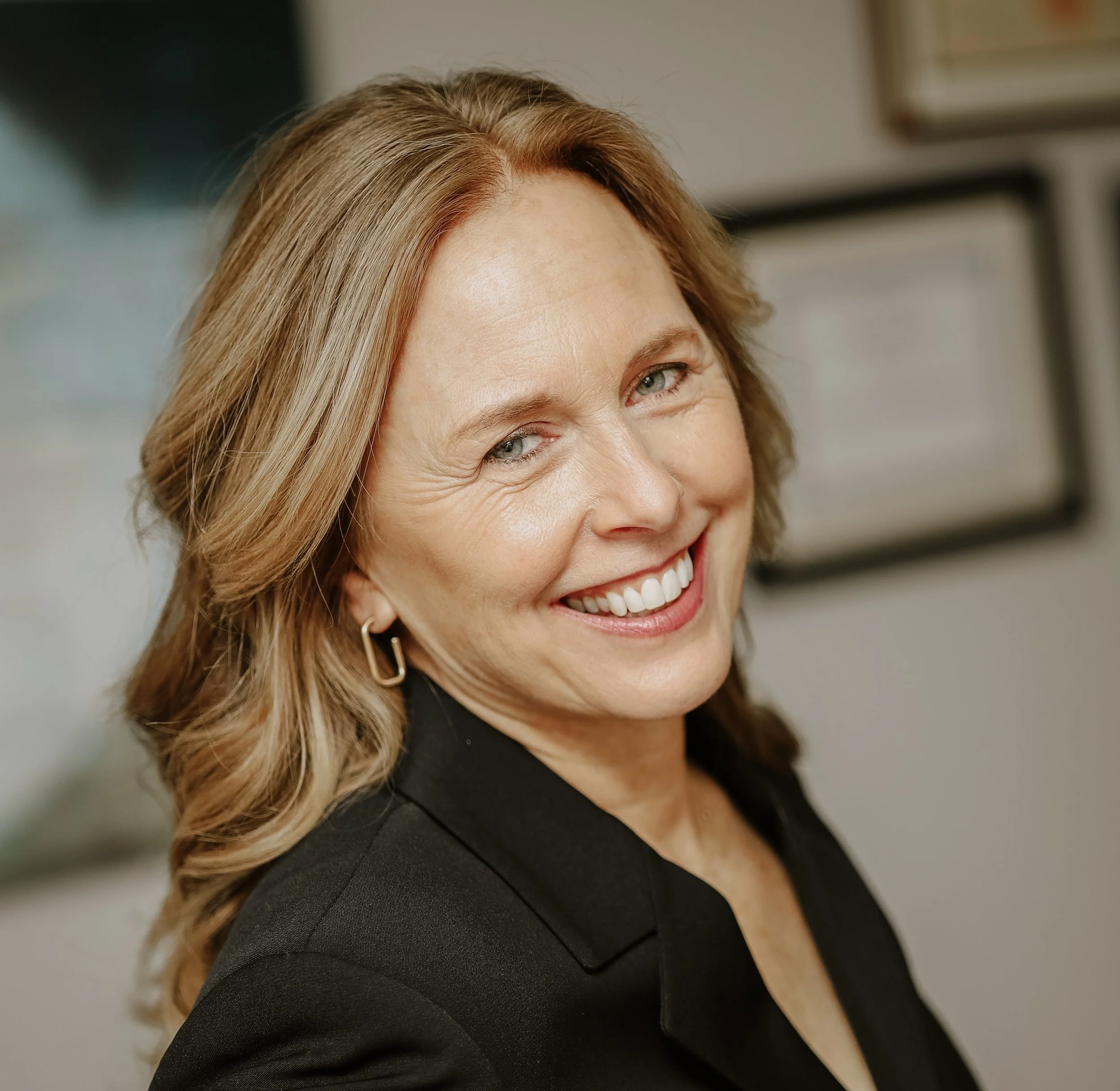 A smiling woman with shoulder-length wavy blonde hair, wearing a black blazer and gold earrings, indoors.