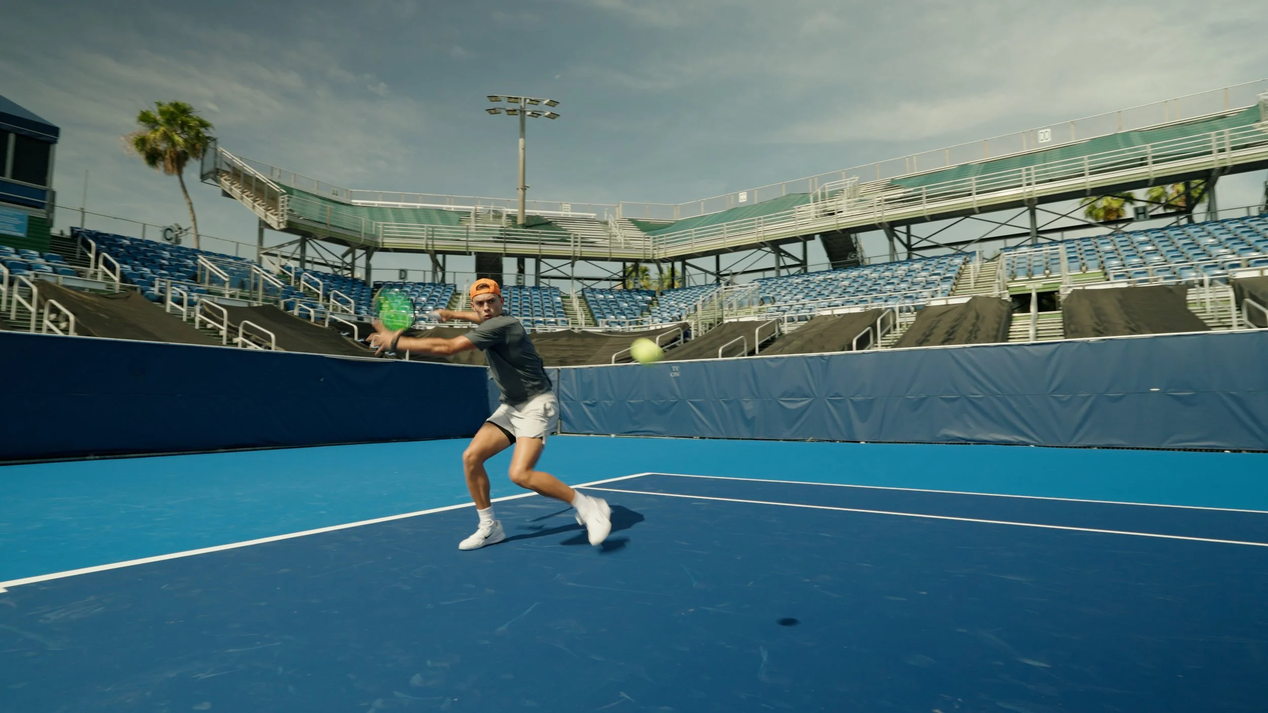 A young male tennis player is preparing to hit a forehand shot on an outdoor tennis court with blue surface, surrounded by empty stadium seating and palm trees under a partly cloudy sky.