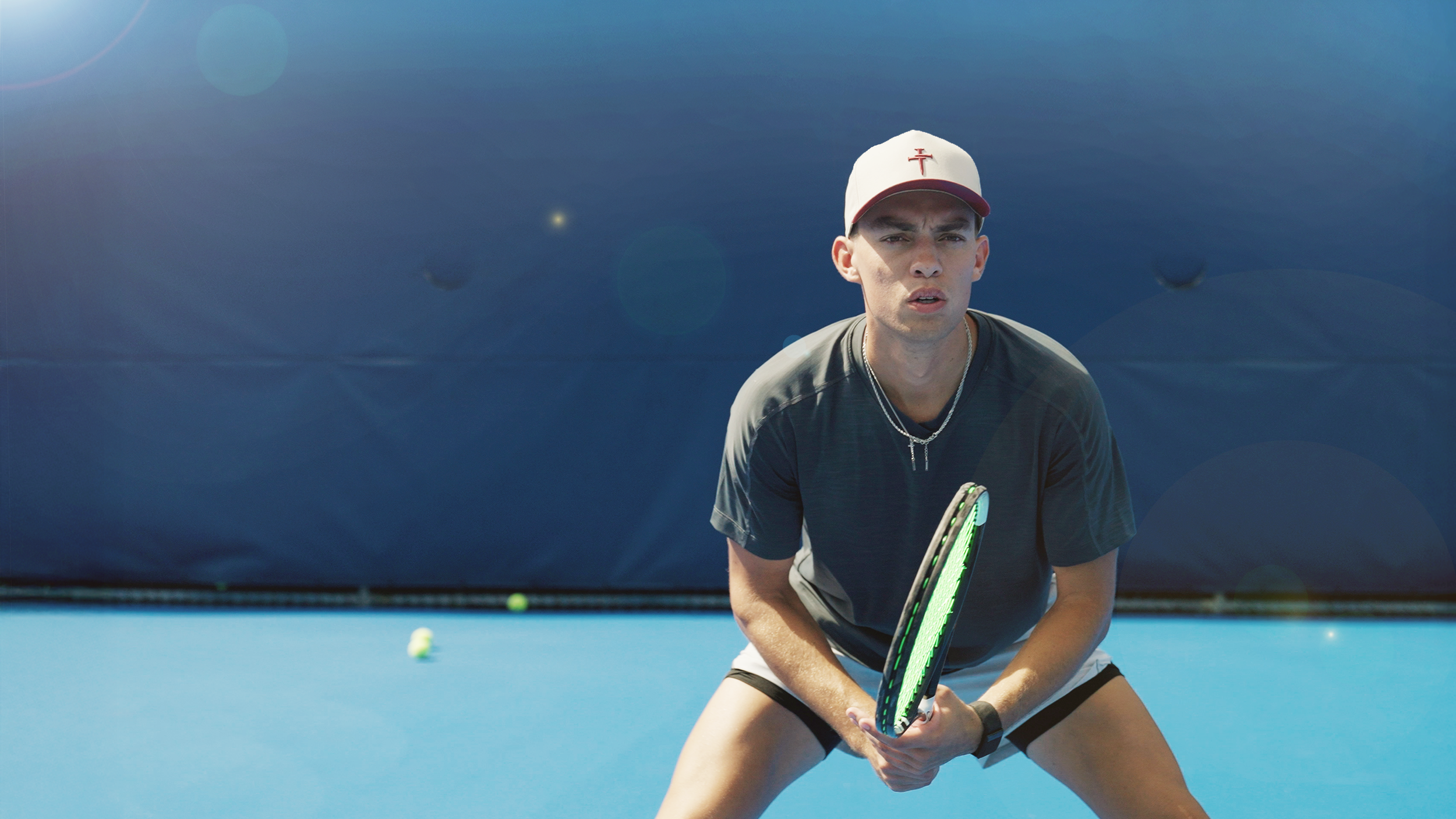 A young man in athletic clothing is playing tennis on an outdoor court, holding a tennis racket, with tennis balls in the background.