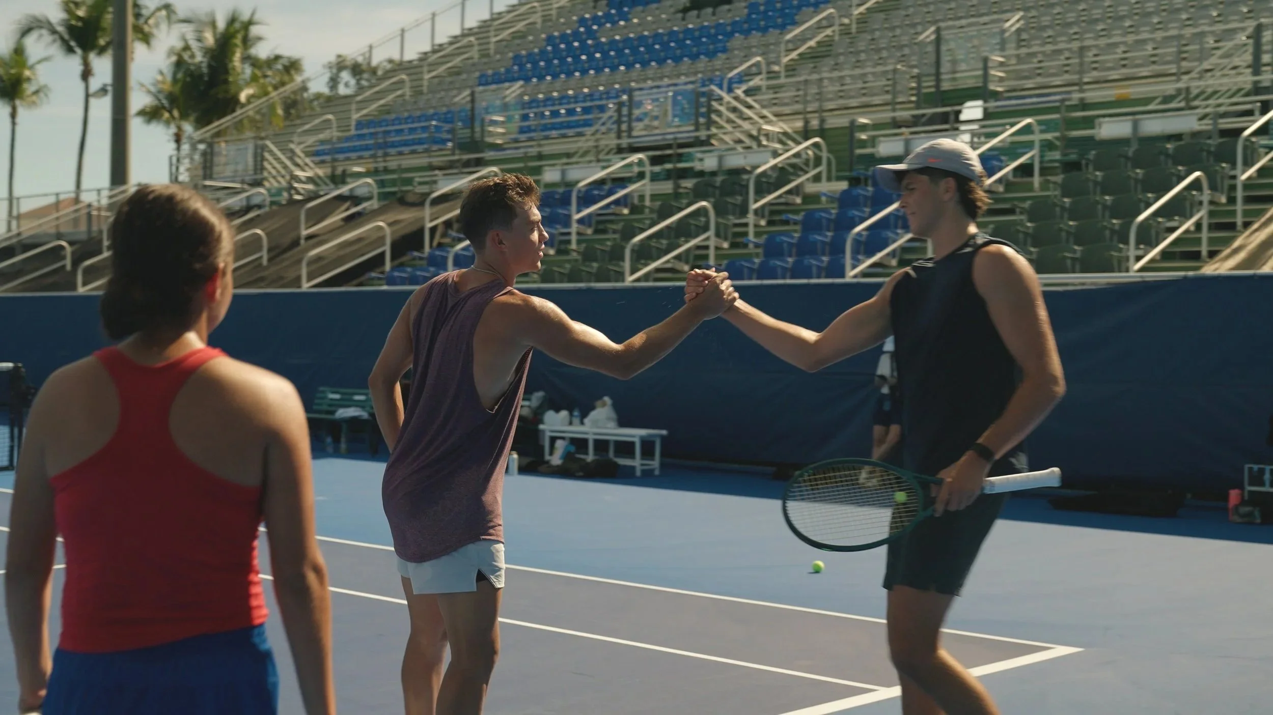 Two male tennis players shaking hands on a tennis court after a match, with a female player watching in the foreground and empty stadium seats and palm trees in the background.