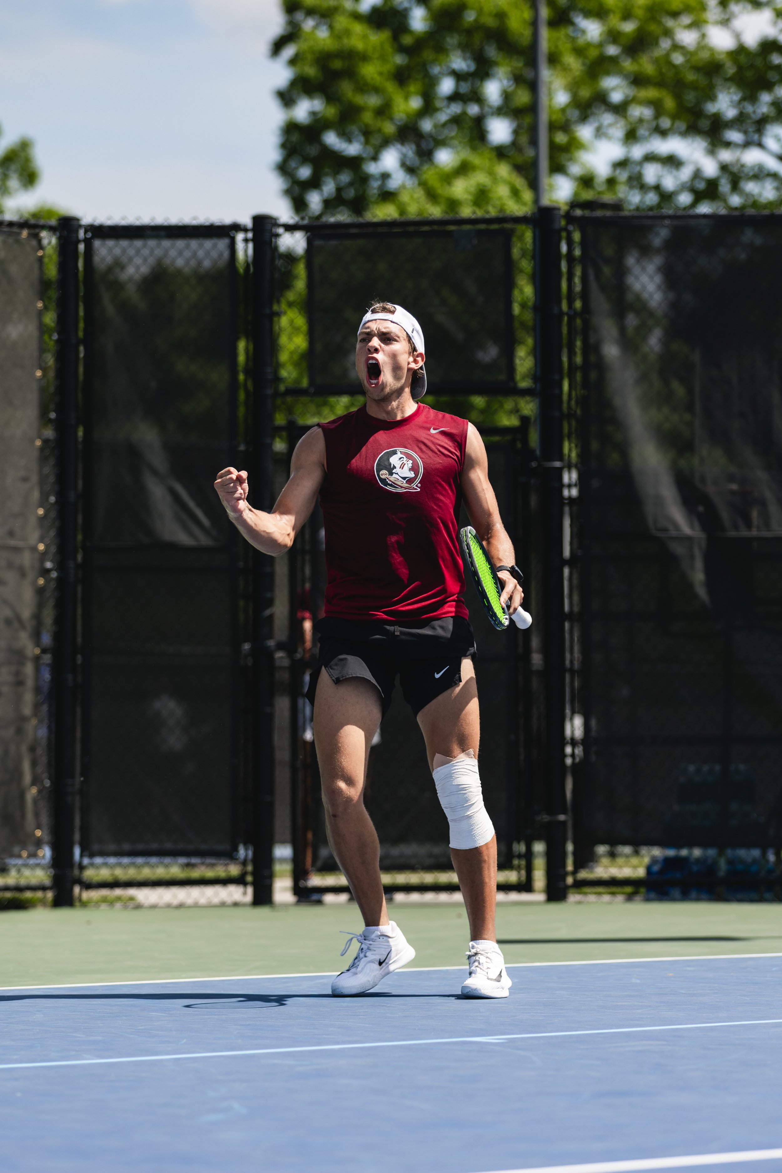 Male tennis player wearing a red sleeveless shirt and white cap celebrating on an outdoor tennis court during the daytime.