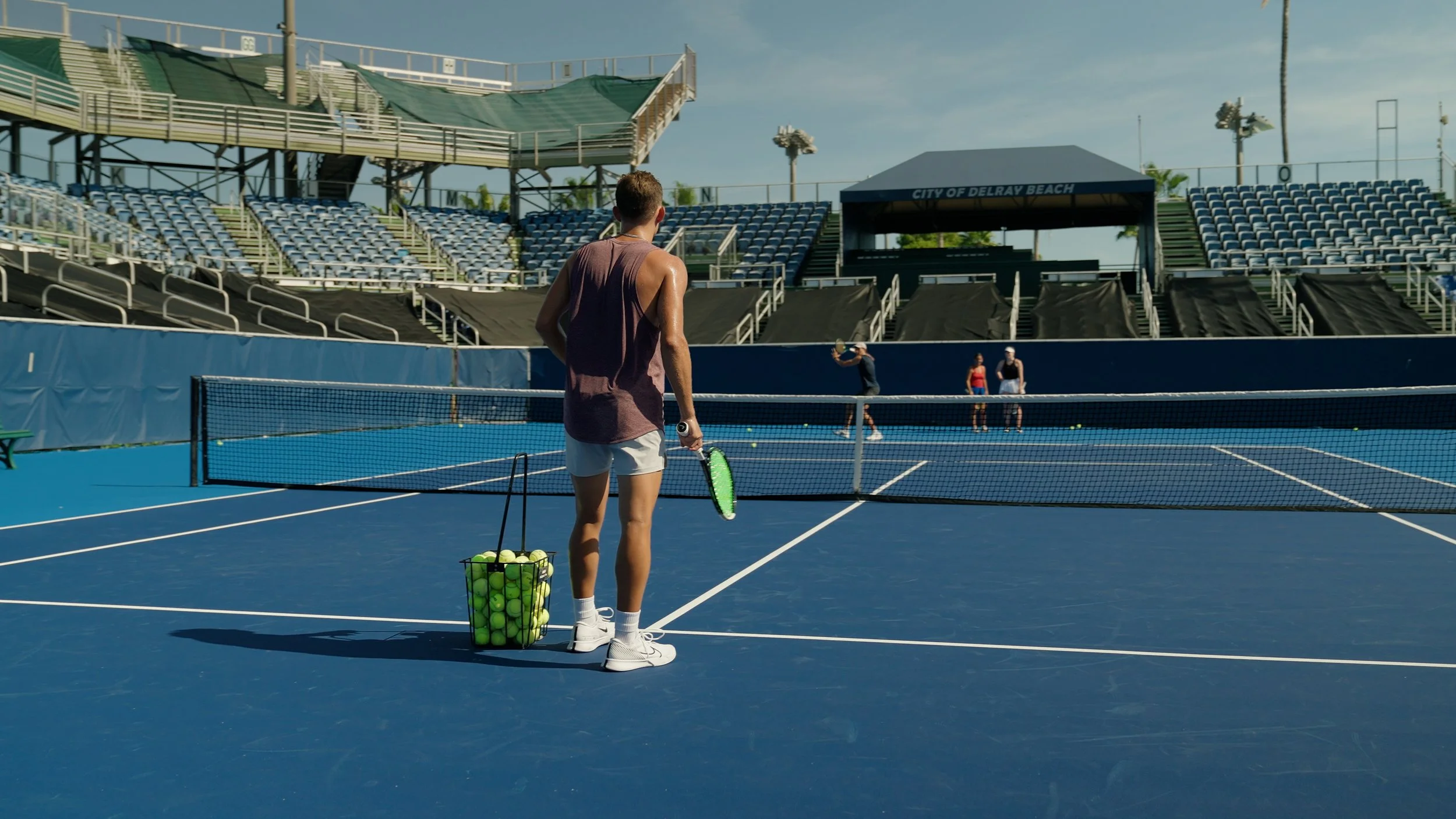 A person standing on a blue tennis court with a basket of tennis balls, holding a tennis racket, preparing for play, with empty stadium seats and a scoreboard in the background.