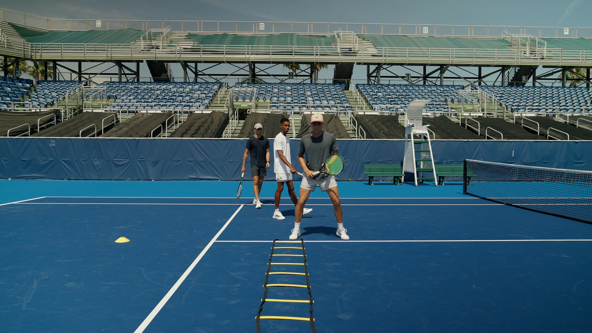 Three young men play tennis on an outdoor court with blue surface, stop training by the net, with bleachers and stadium structure in the background.