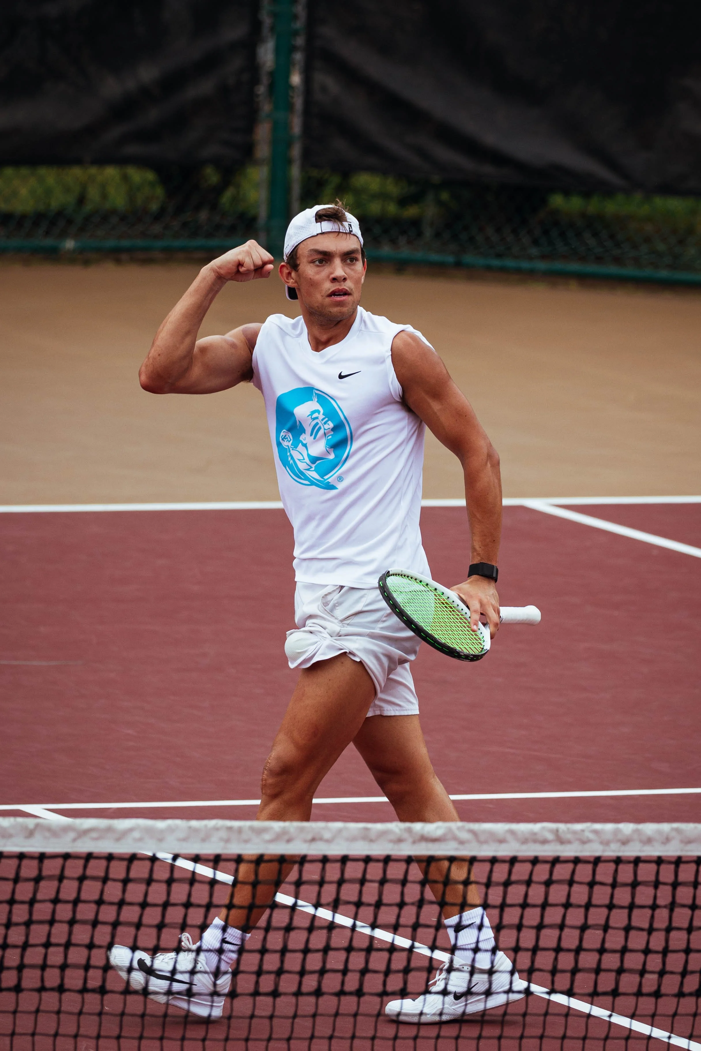 A male tennis player in white sportswear, including a white cap worn backwards, is standing on a tennis court near the net. He is holding a tennis racket in his right hand and flexing his bicep with his left arm.