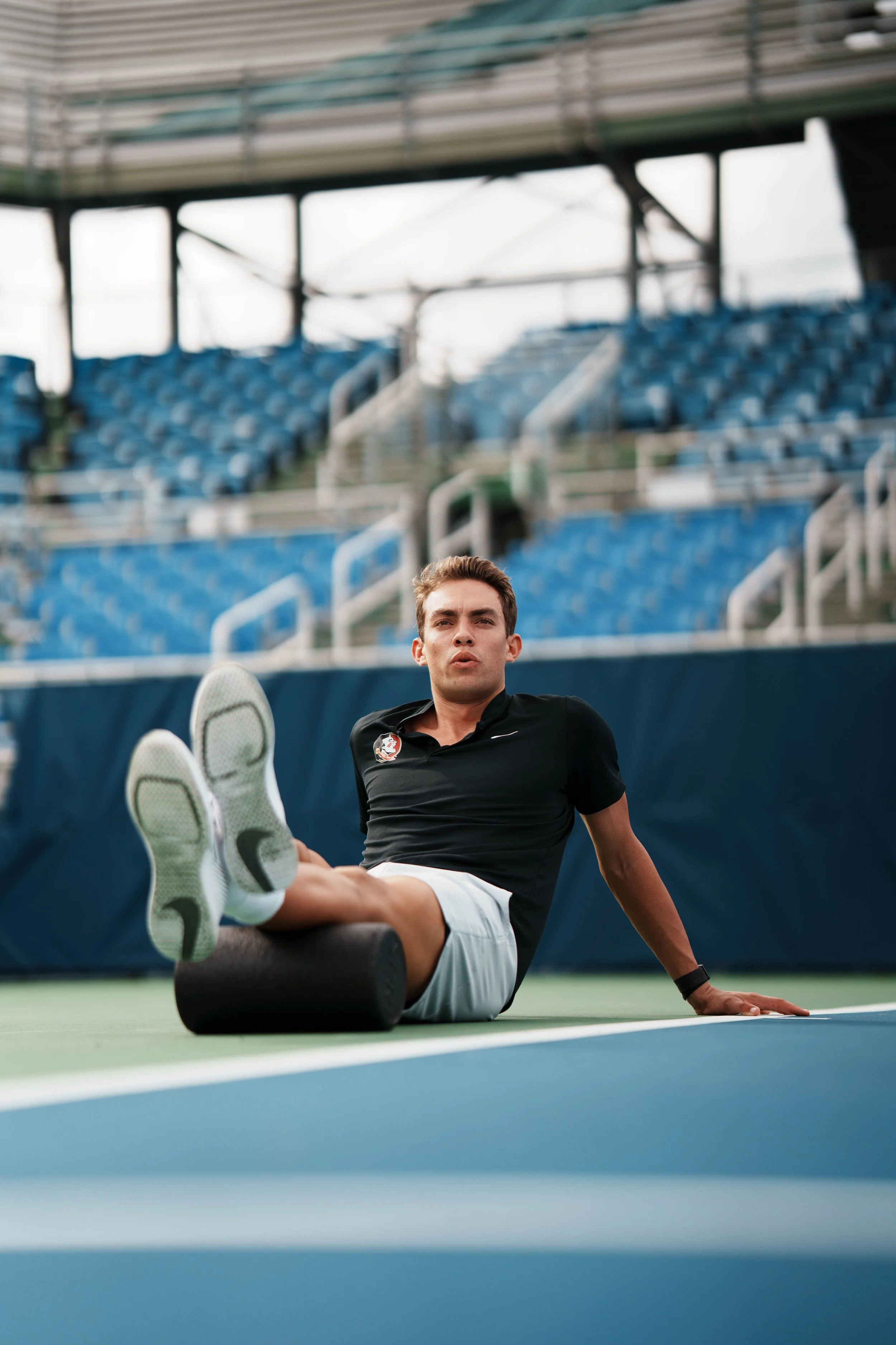 A man sits on a tennis court with his legs extended, wearing a black shirt and gray shorts, with a foam roller under his leg, during daytime. Empty stadium seats are visible in the background.