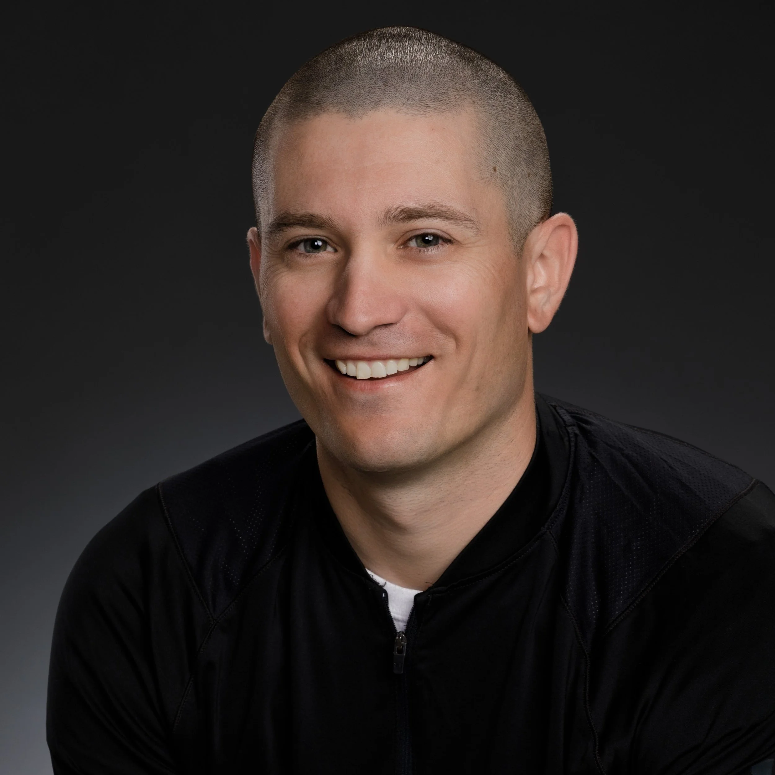 Close-up of a smiling man with short hair wearing a black jacket and white shirt against a dark background.
