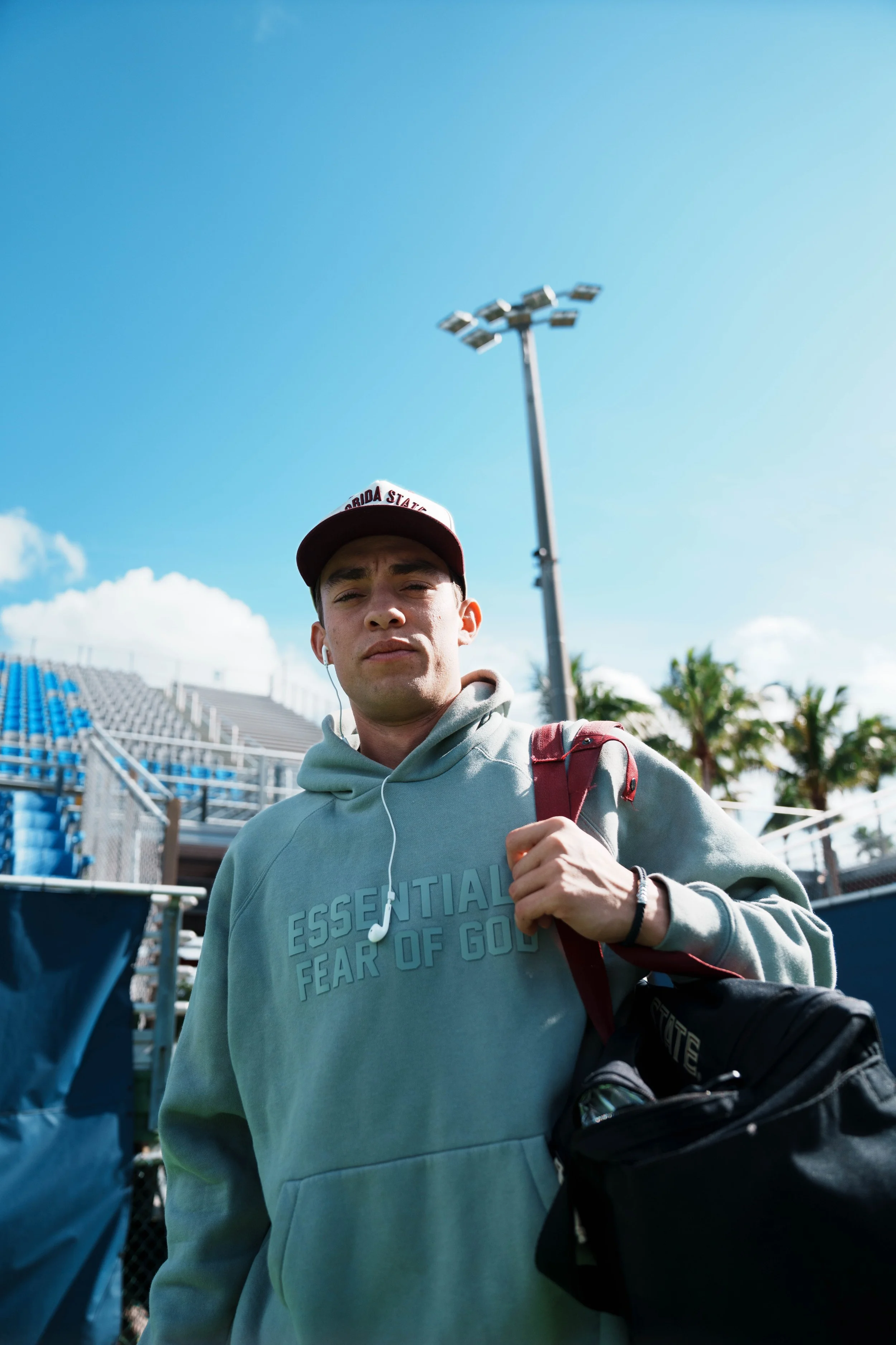 Young man wearing a gray hoodie with text, a red and white cap, and earphones, carrying a black bag over his shoulder, walking outdoors near stadium bleachers and palm trees on a sunny day.