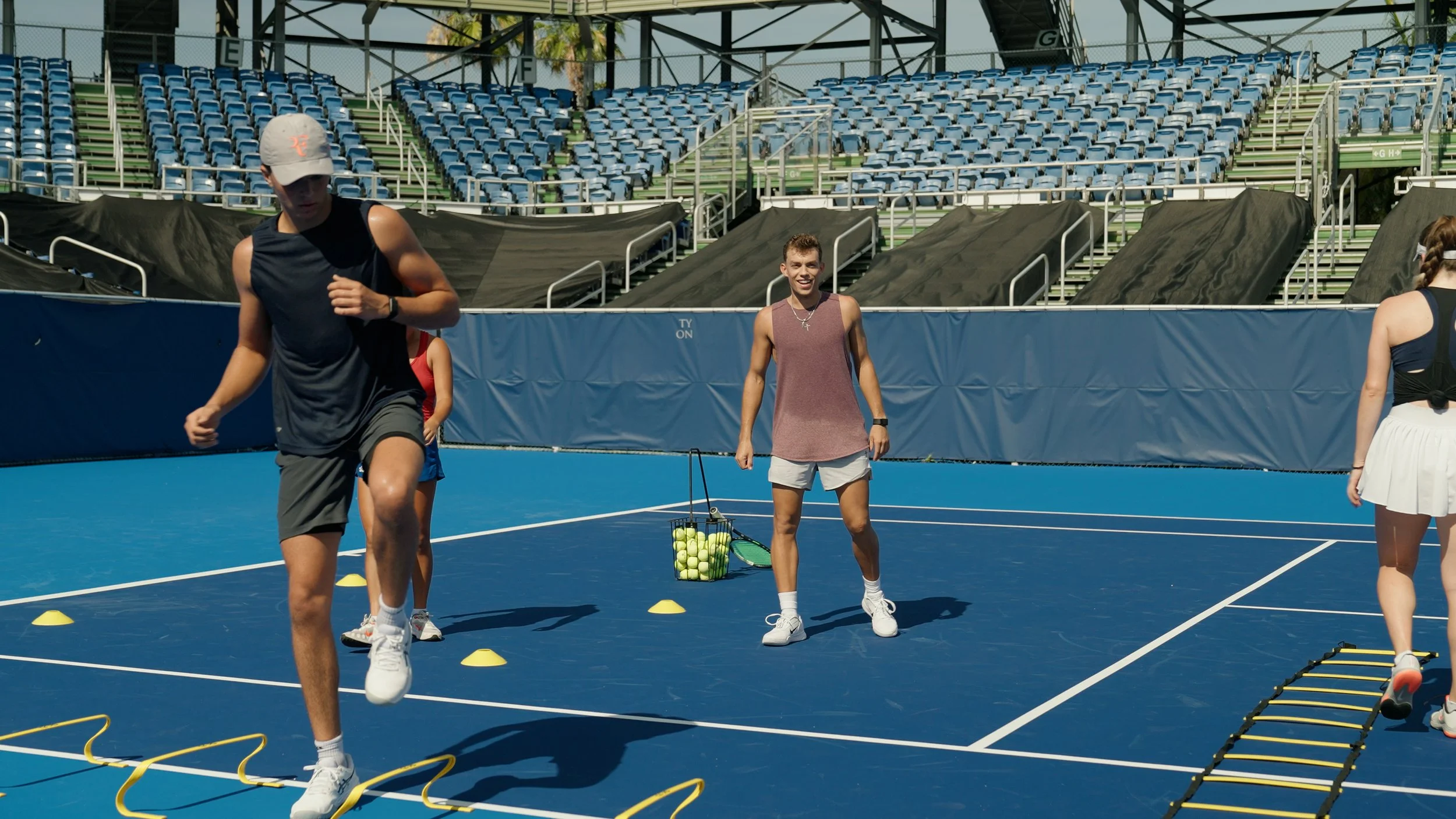 People practicing tennis drills on a blue tennis court at an outdoor stadium with empty blue seats in the background.