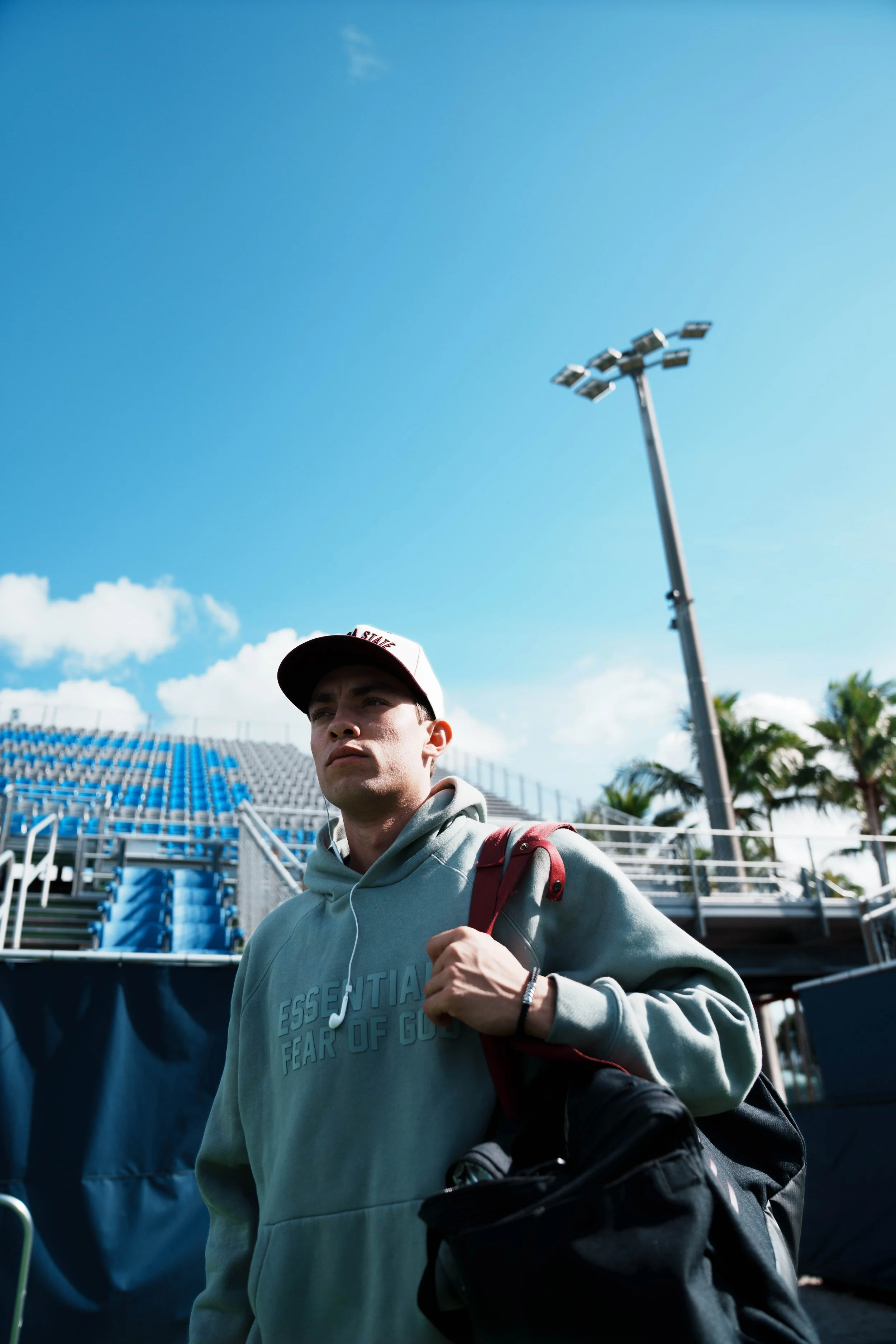 A young man wearing a gray hoodie with the words 'ESSENTIAL FEAR OF GOD' on it, carrying a red backpack, walking past stadium seats and metal bleachers, with a tall stadium light pole and blue sky in the background.