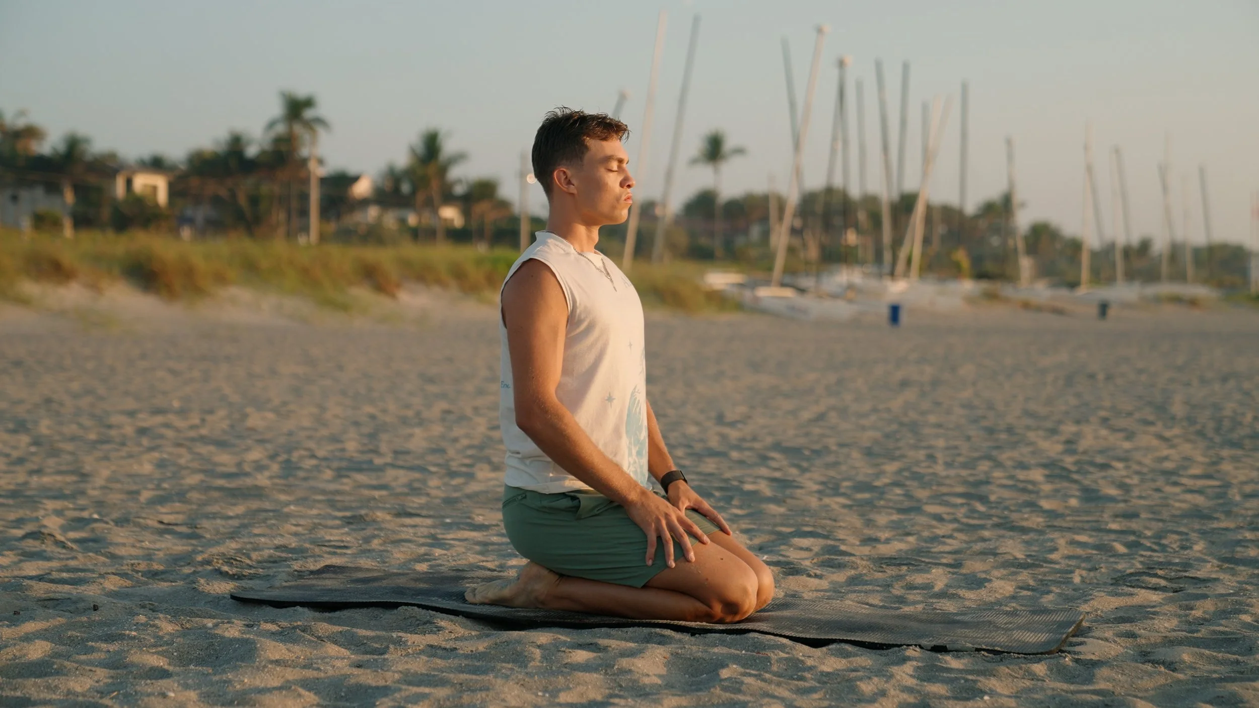 A young man kneeling on a yoga mat on a sandy beach, with his hands resting on his thighs, eyes closed, appearing to meditate during sunset, with sailboats and palm trees in the background.